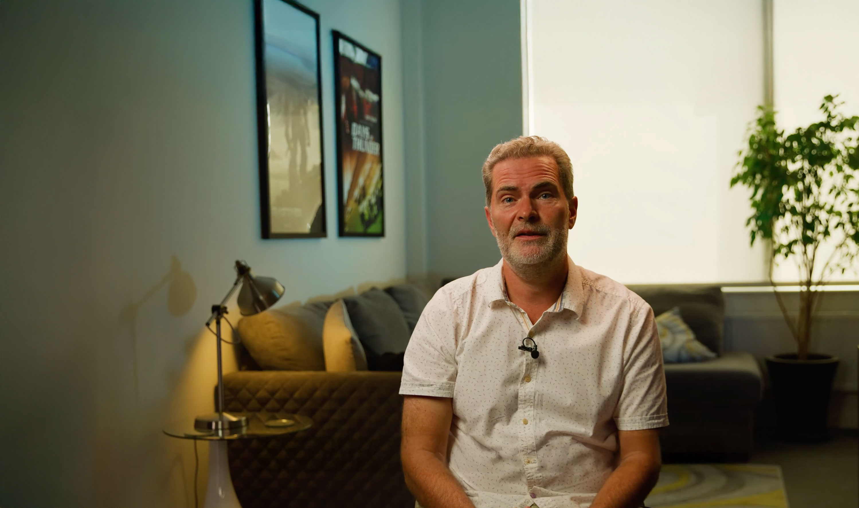 A man in a white shirt sits in a living room with framed posters on the wall.