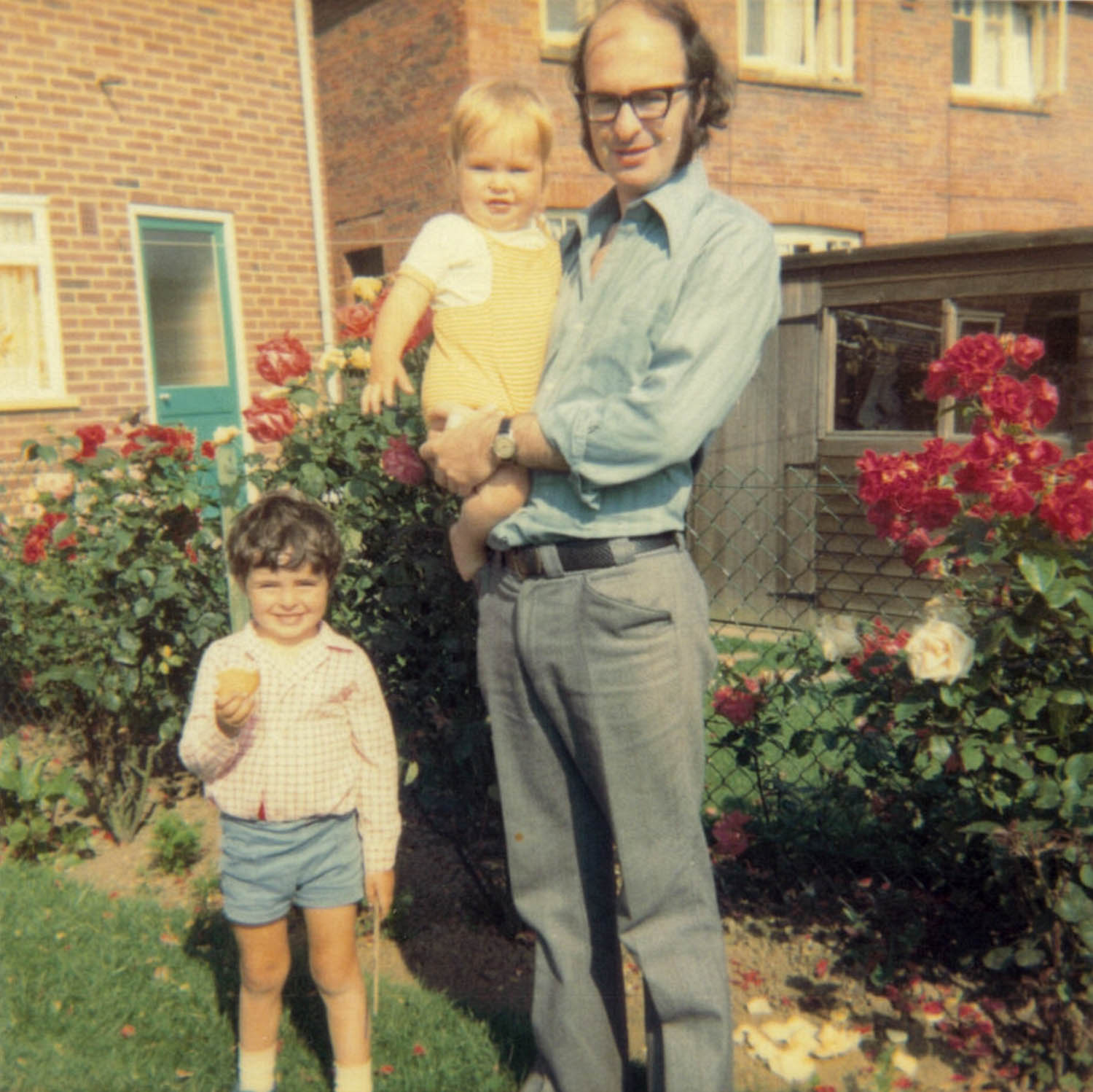 A man holds a child while another child stands beside him in a garden with roses.