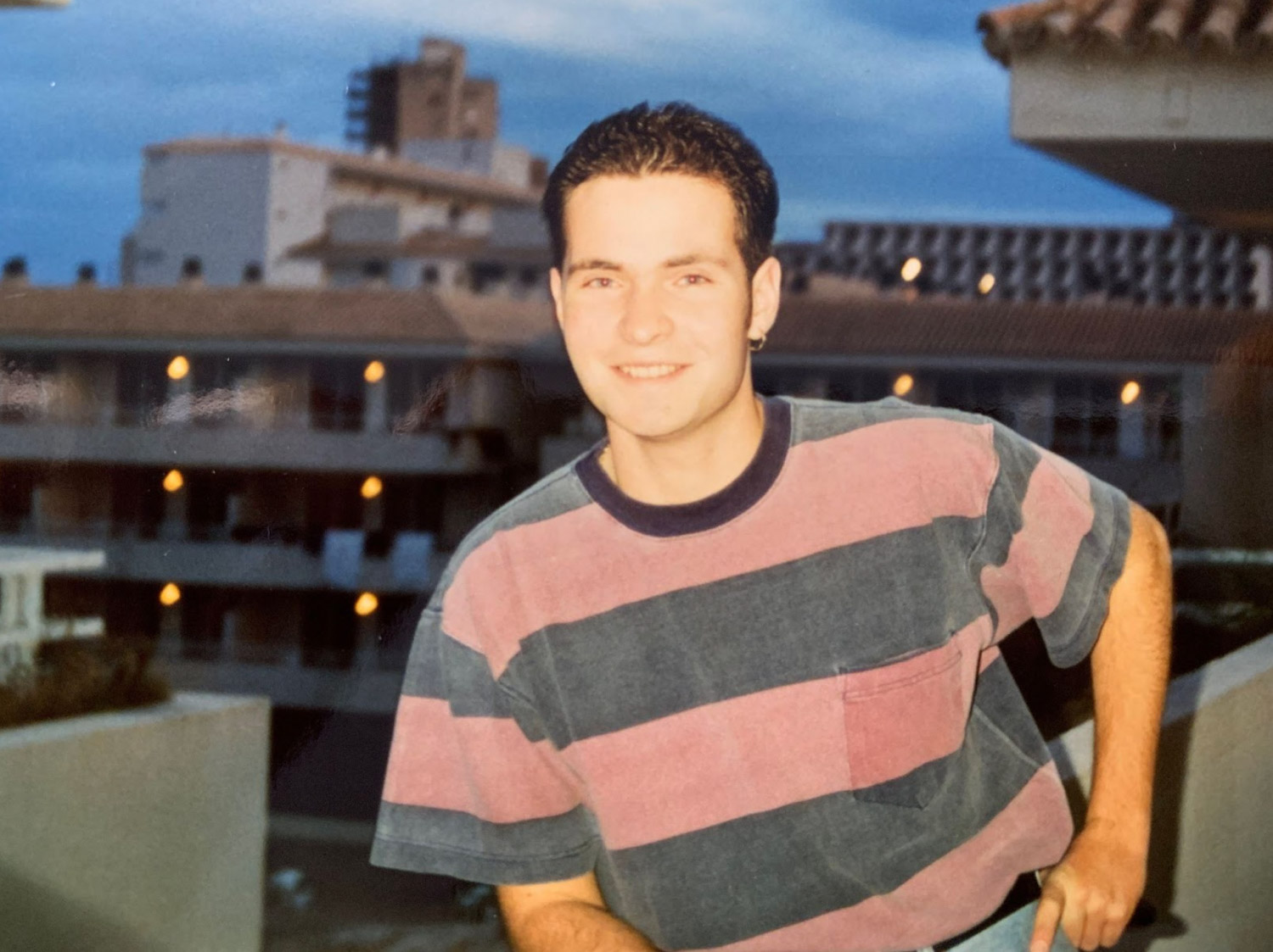 Young man smiling and leaning against balcony railing. Urban buildings are in the background.
