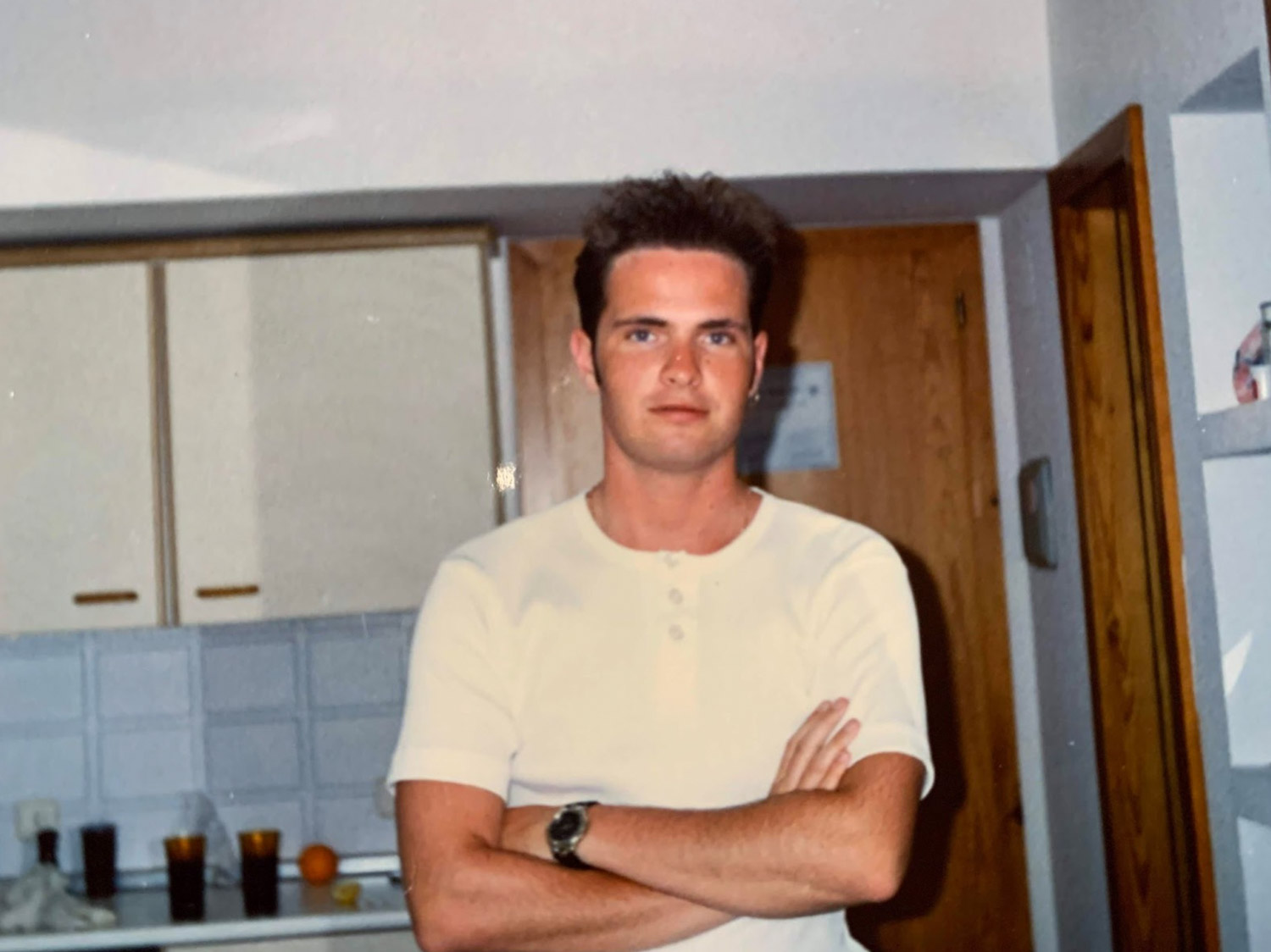 A person in a white shirt stands with arms crossed in a kitchen area.