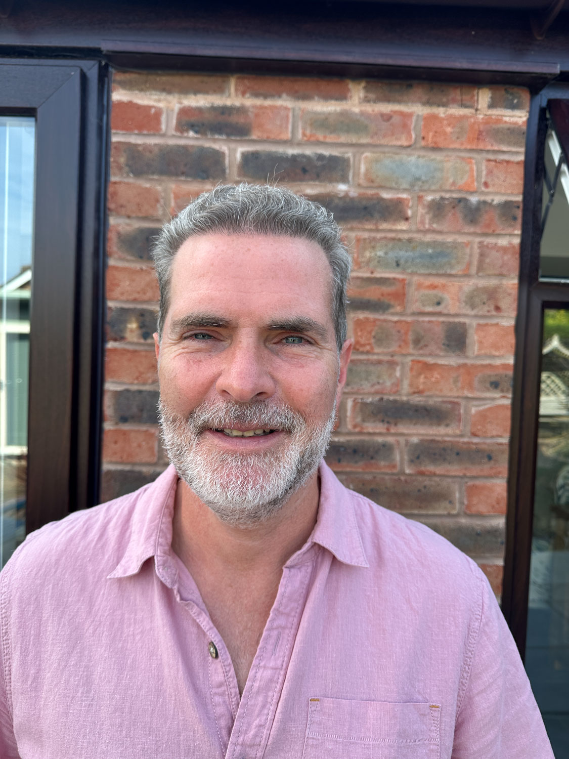 A man with short hair and a beard smiles, standing in front of a brick wall.