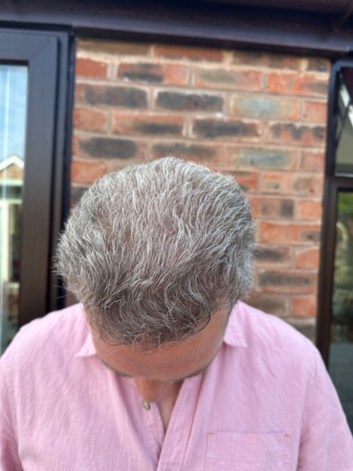 A person in a pink shirt looks down, standing in front of a brick wall outside.