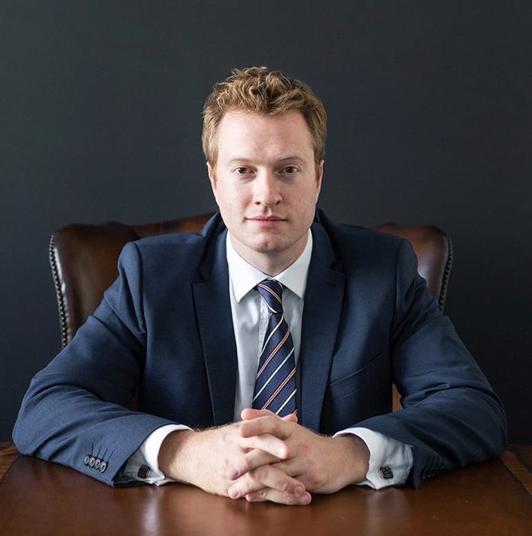 A man in a suit sits at a desk with hands clasped, looking forward.