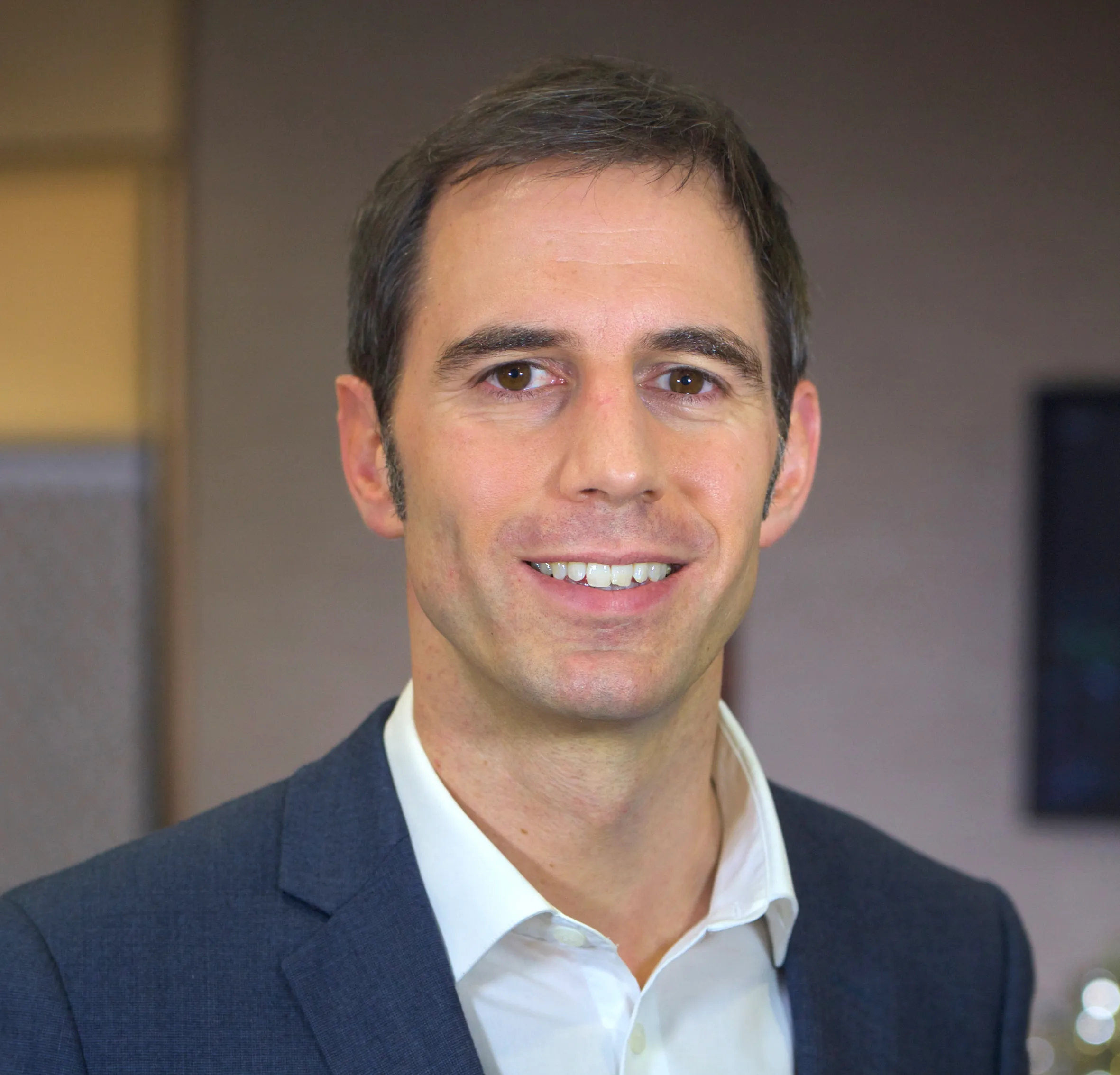 A man in a suit smiles directly at the camera in an office setting.