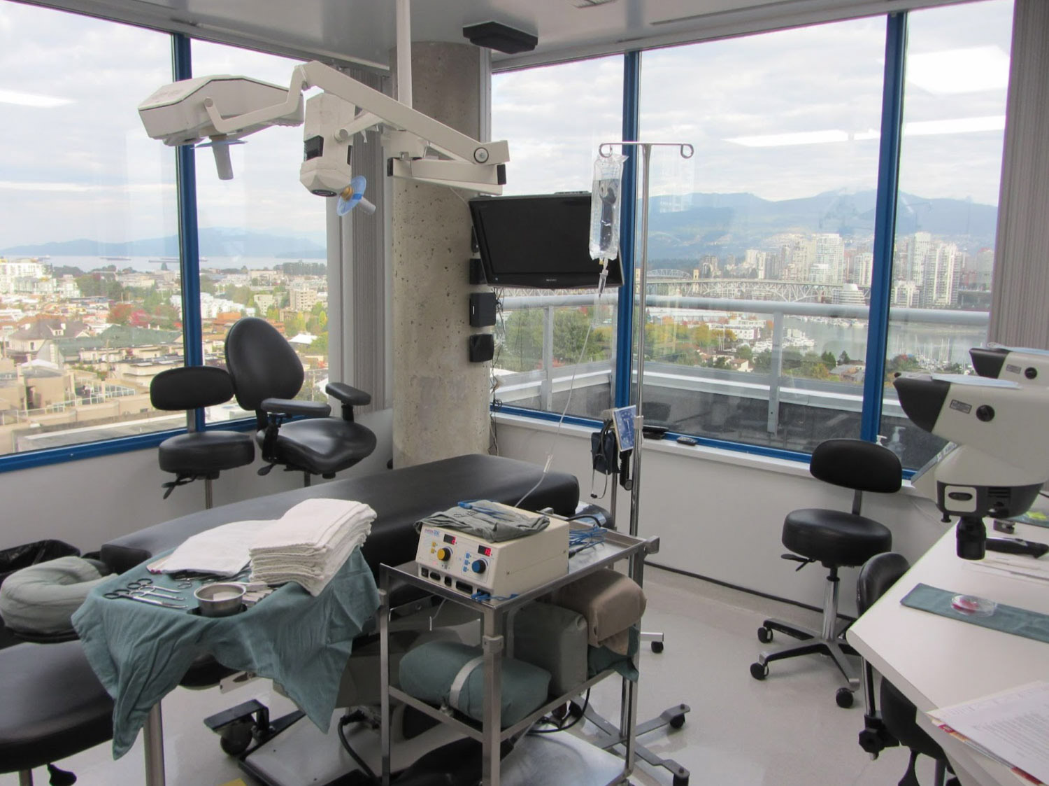 A clean, modern surgical room with operating table, medical equipment, and large windows with city view.