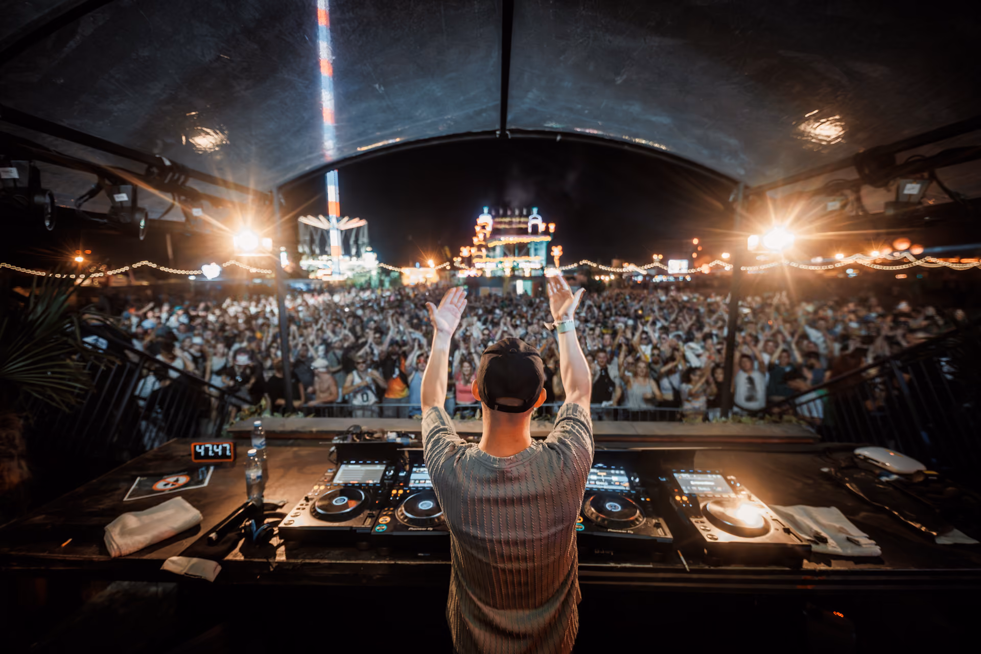 DJ wearing a black cap and a gray shirt stands at the mixing console with hands raised, facing a large outdoor crowd at night with festival lights and structures in the background.