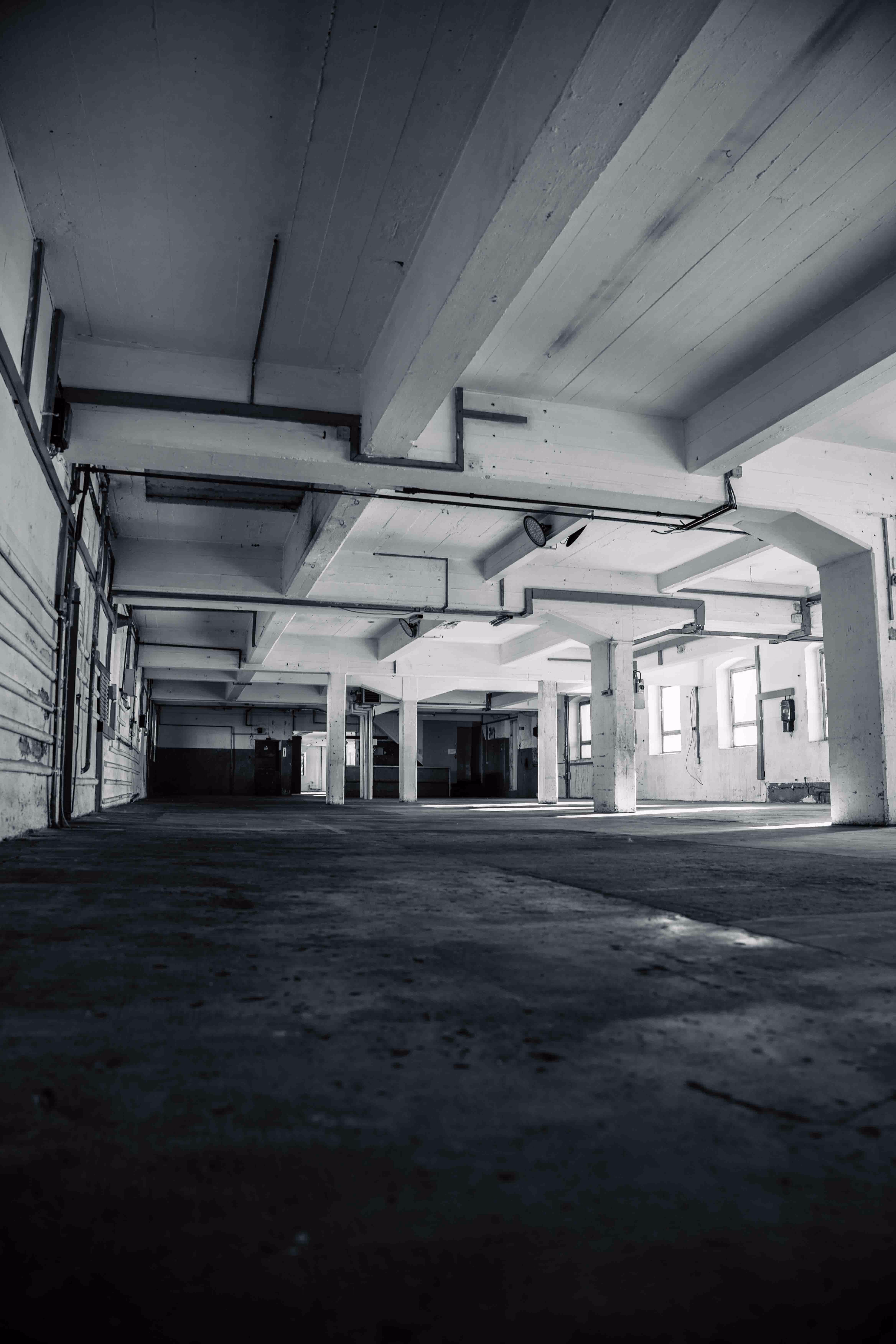 Interior of an empty industrial warehouse with white concrete pillars and beams, large windows letting in light, and a dark concrete floor.