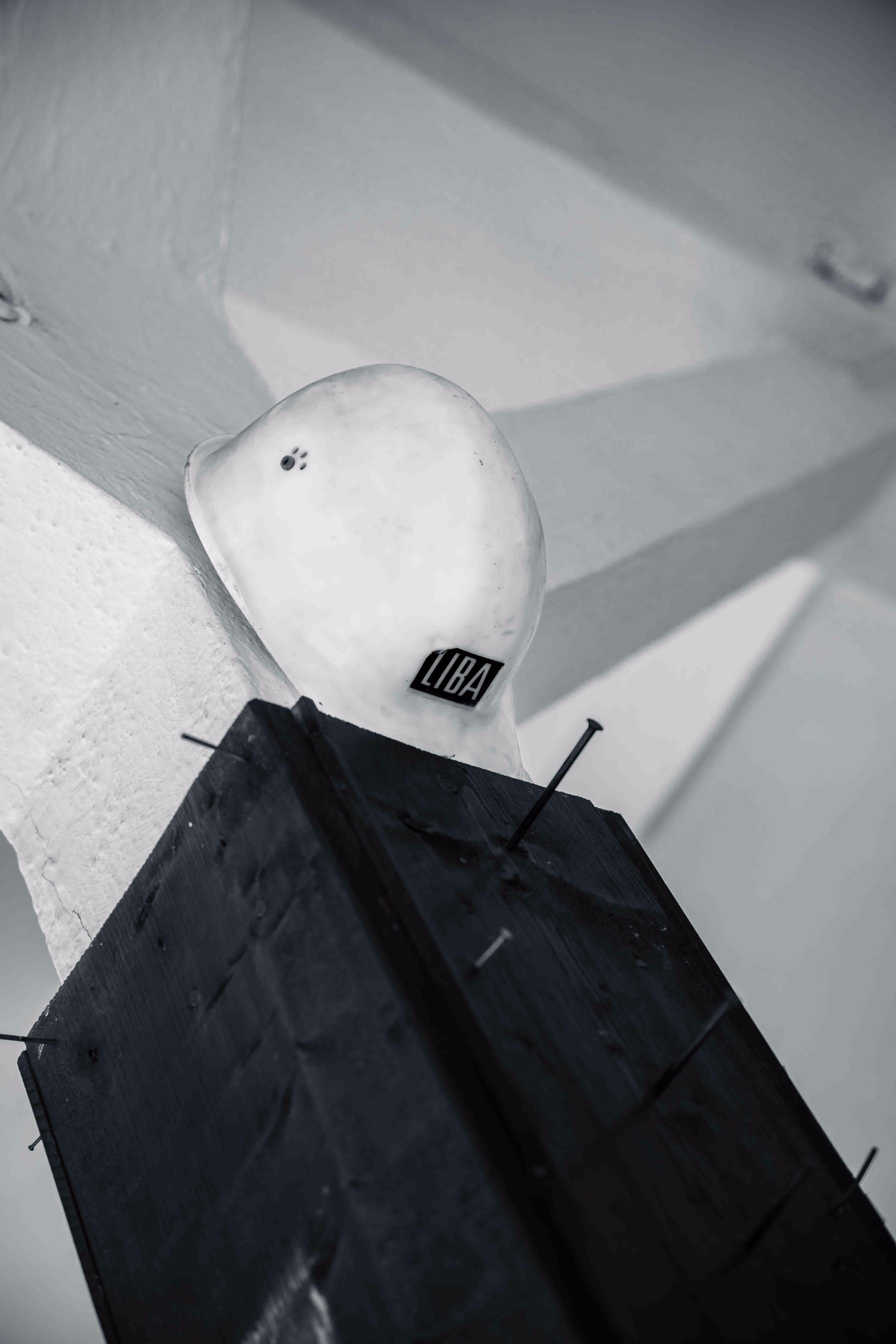 White construction helmet with a 'LIBA' label placed on top of a tall black rectangular structure with protruding nails, set against a light, shadowed wall background.