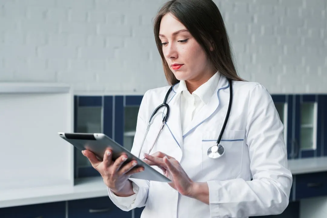 A female doctor in a white coat reviews patient data on a tablet.