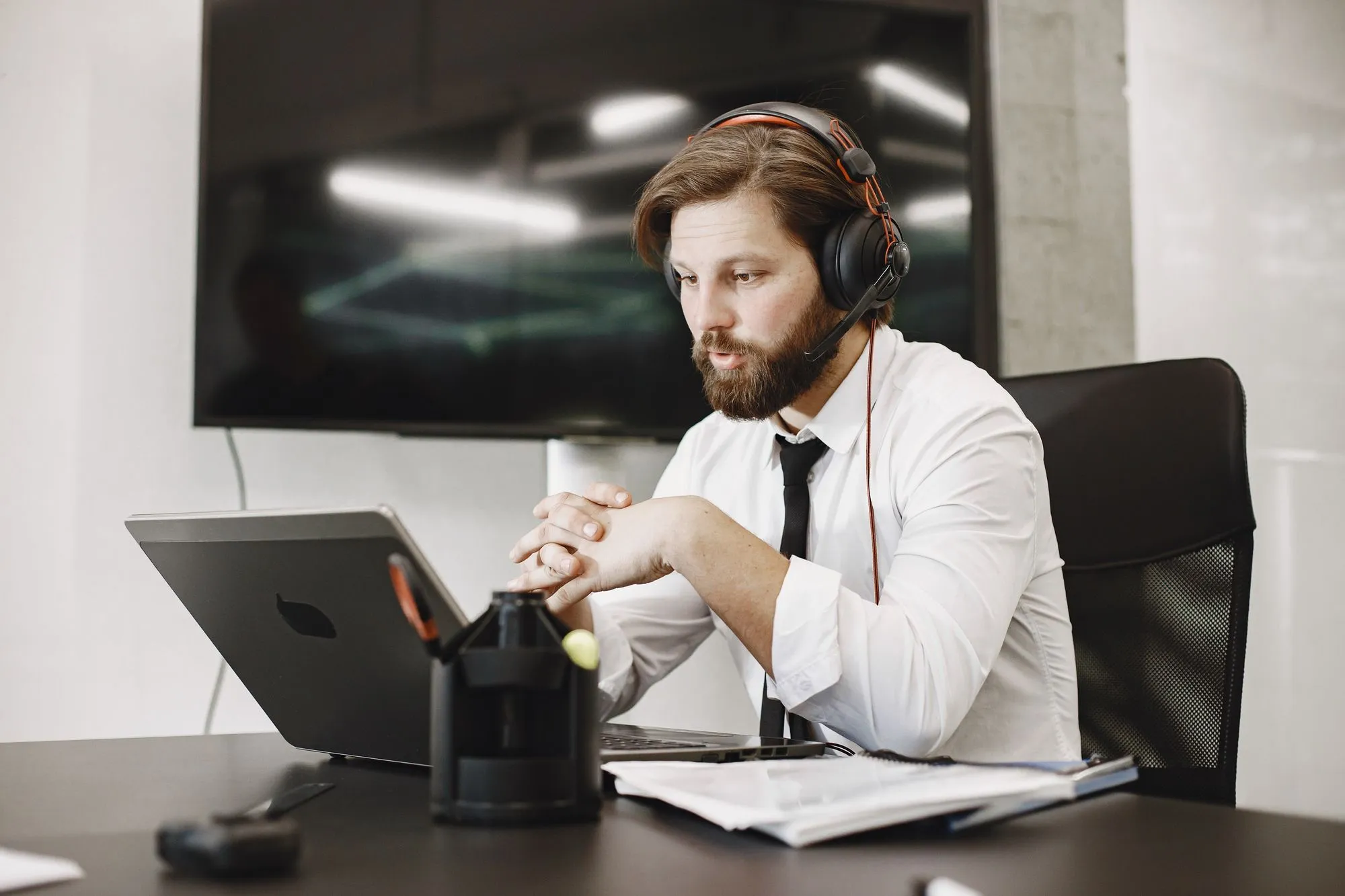 A bearded man wearing headphones sits at a desk with a laptop, focused on music.