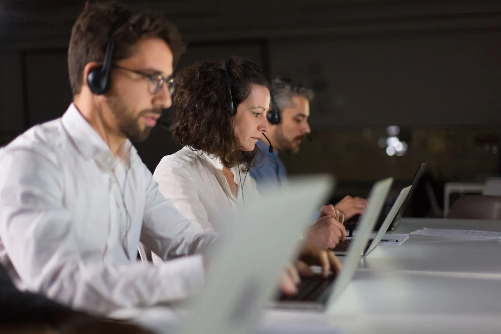 Three Customer services employees wearing headsets, engaged in conversation while seated at their desks.