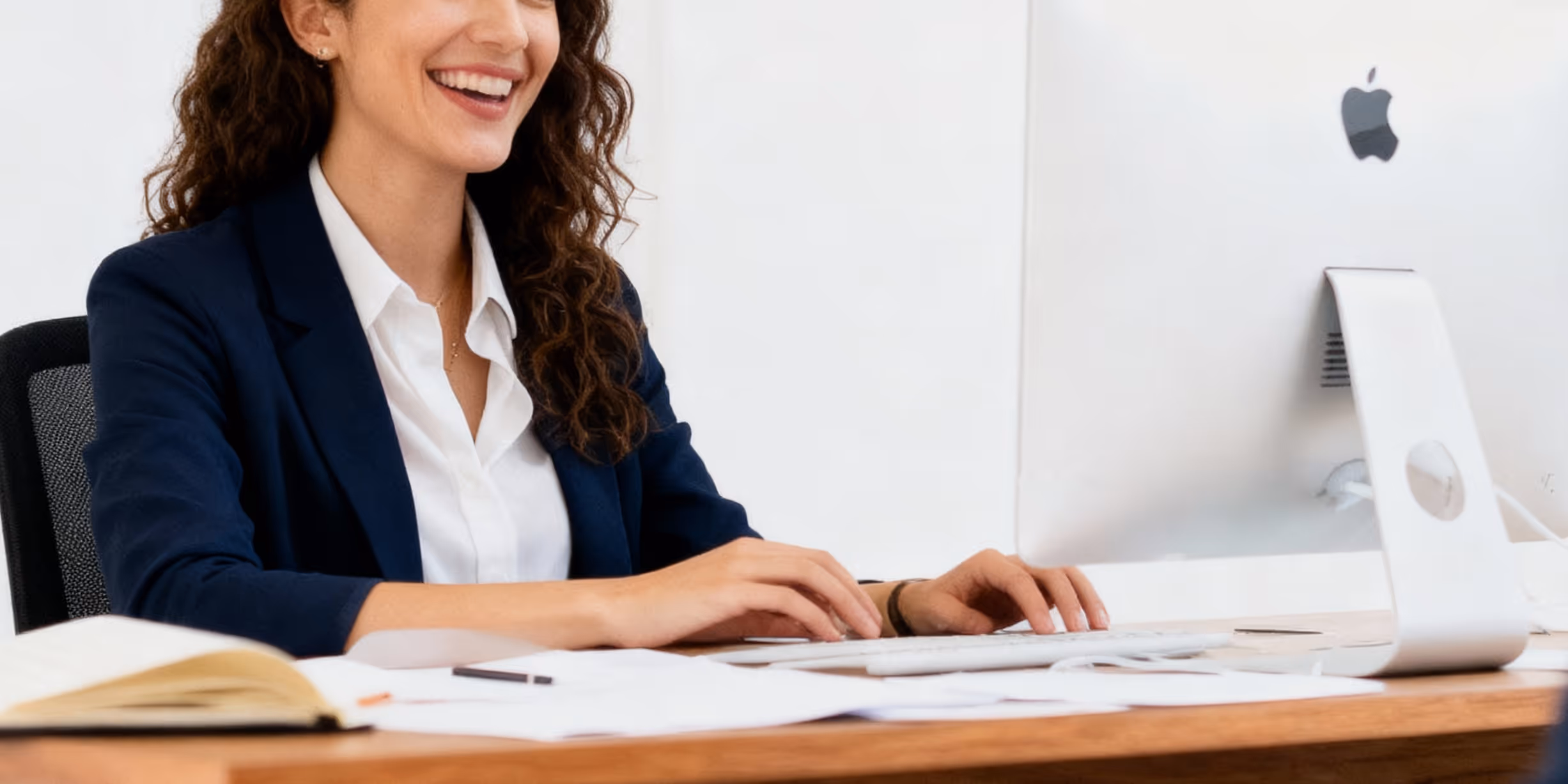 Smiling woman in a white shirt and navy blazer typing on a white keyboard at a desk with papers and an open book, using an Apple desktop computer.