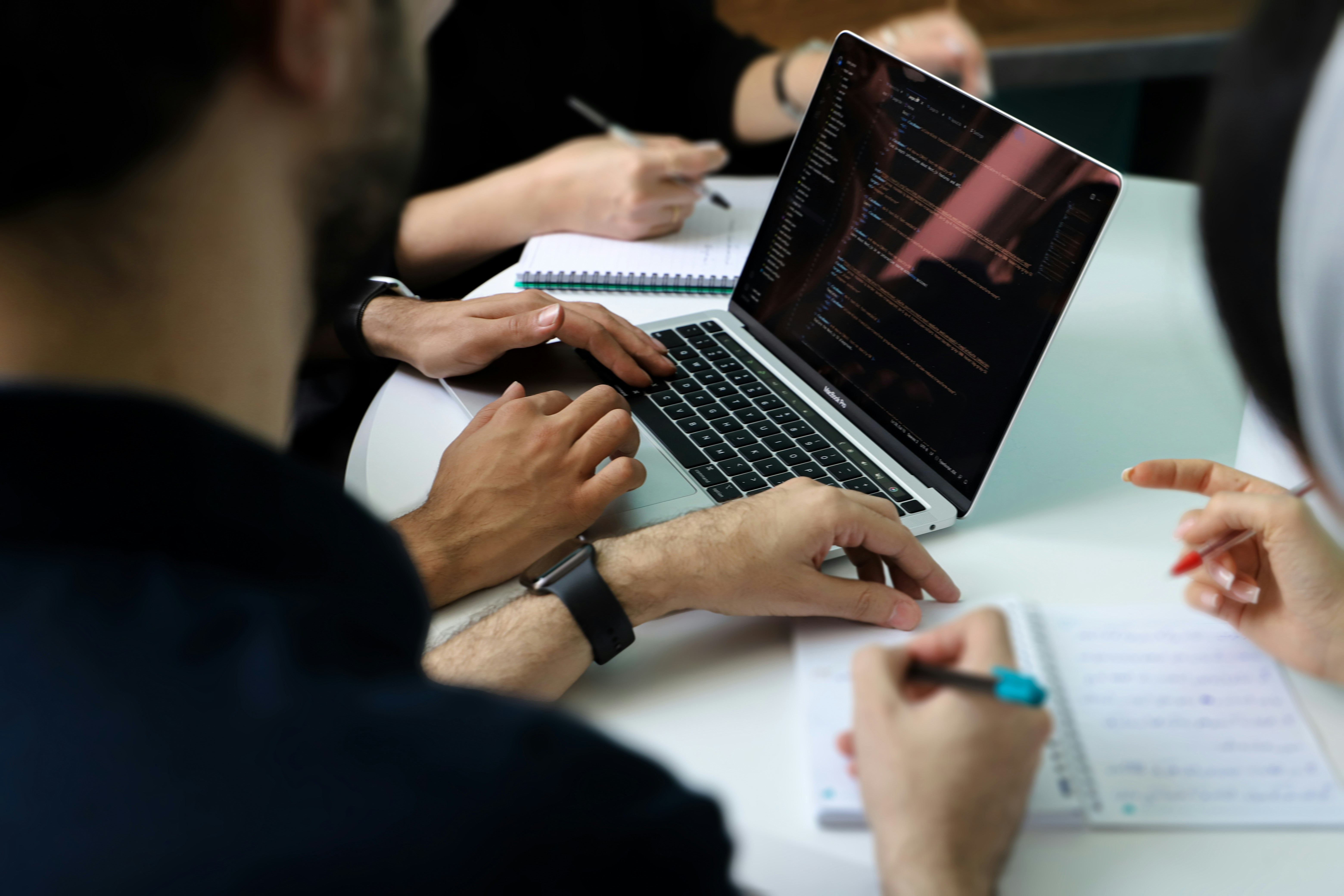 A group of people work together at a desk, referring to a laptop and taking notes.