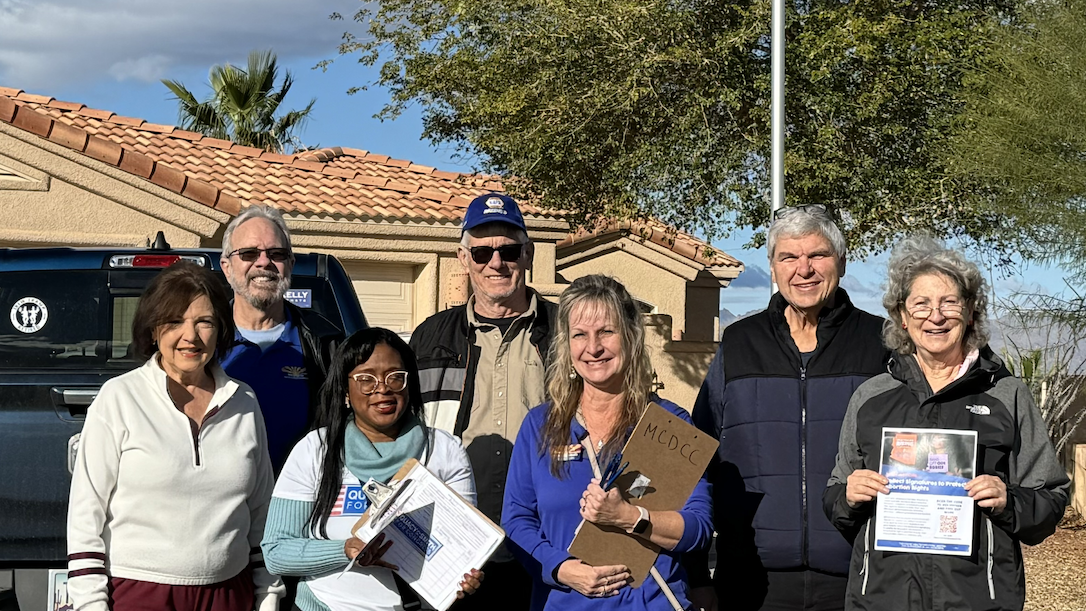 Group of people outdoors holding clipboards and flyers, facing the camera and smiling.