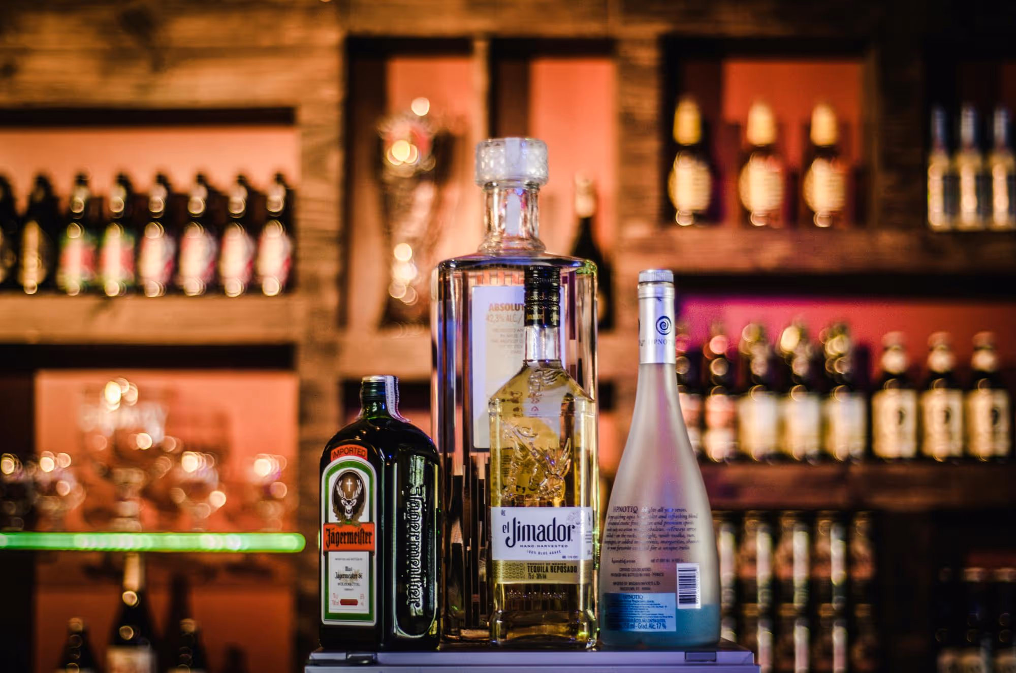 Four liquor bottles including Jägermeister, El Jimador tequila, Absolut vodka, and Hypnotiq on a bar counter with shelves of bottles in the background.