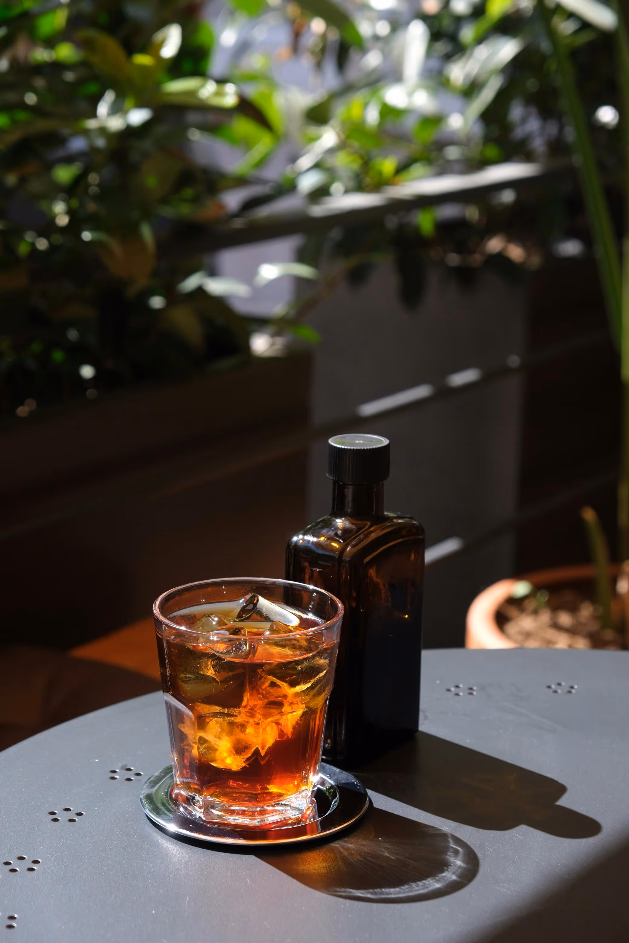 Glass of iced amber-colored drink on a metal coaster beside a dark brown bottle on a gray table with plants in the background.