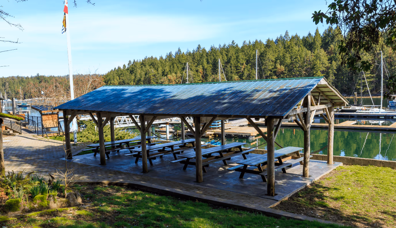 Covered wooden picnic shelter with benches near a marina surrounded by trees and boats.