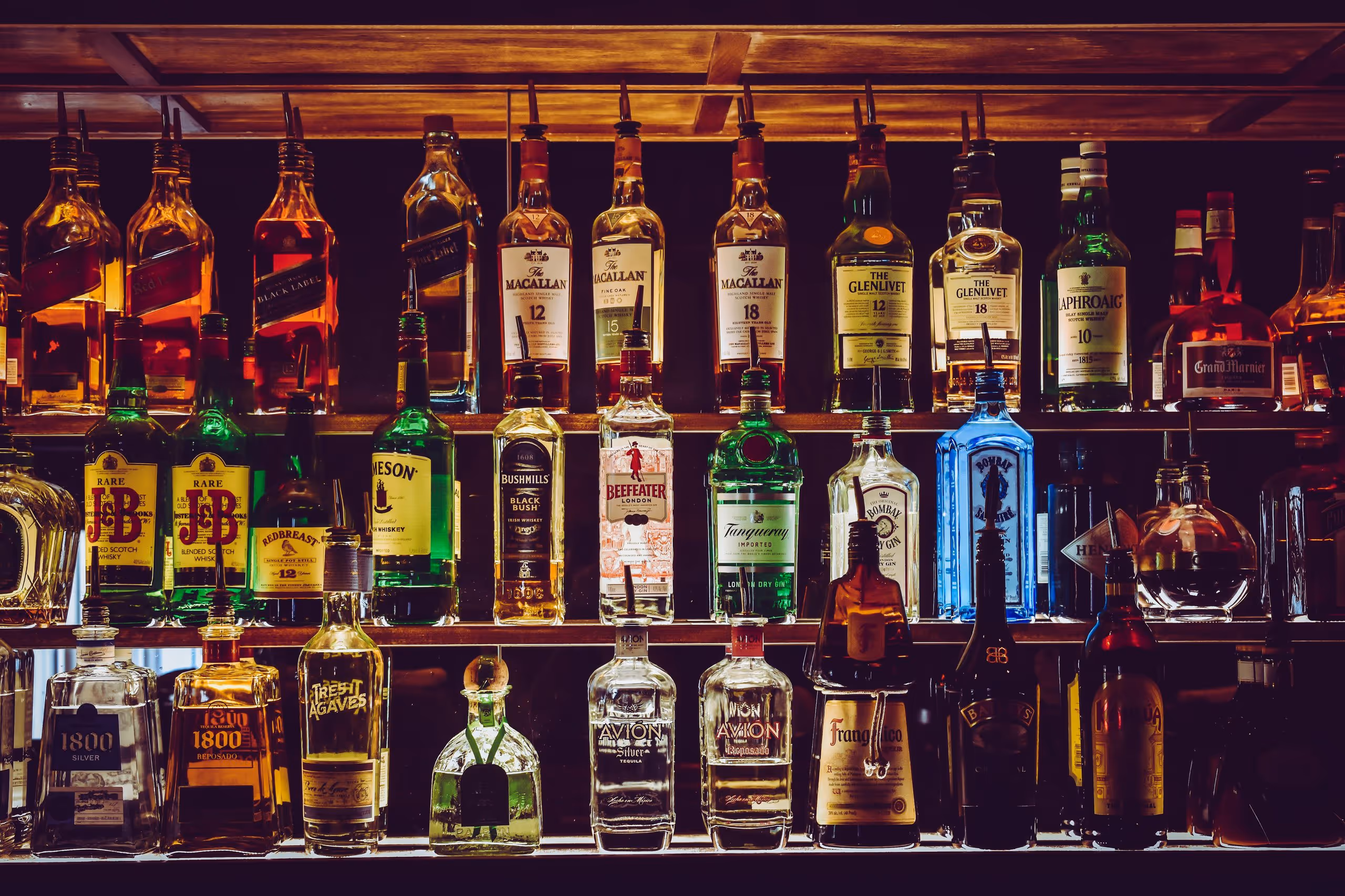 Shelf of various whiskey and liquor bottles including The Macallan, Glenlivet, Laphroaig, Jameson, Tanqueray, and Avion tequila in a dimly lit bar.