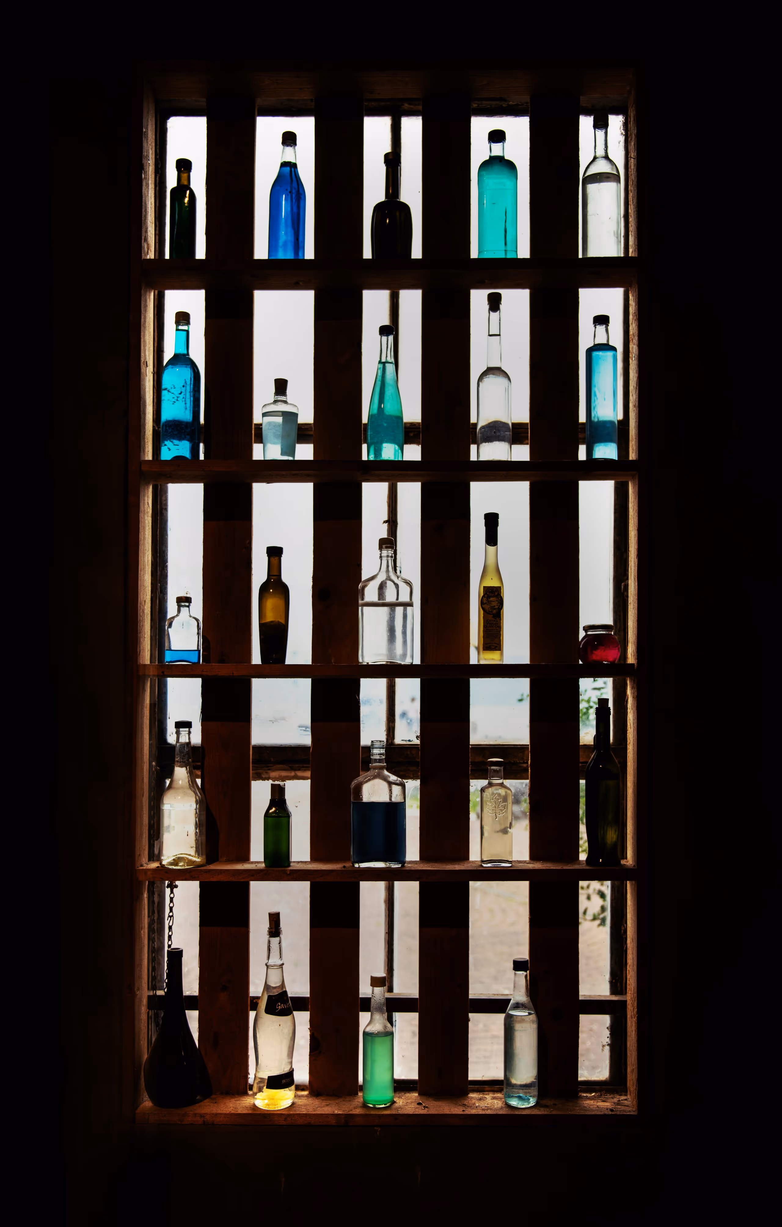 Wooden window shelf holding various glass bottles filled with clear and colored liquids, backlit by daylight.