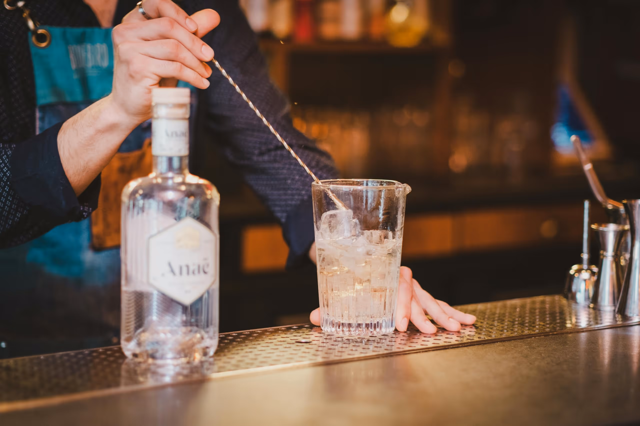 Bartender stirring a clear drink with ice in a mixing glass on a bar counter next to a bottle labeled Anaé.