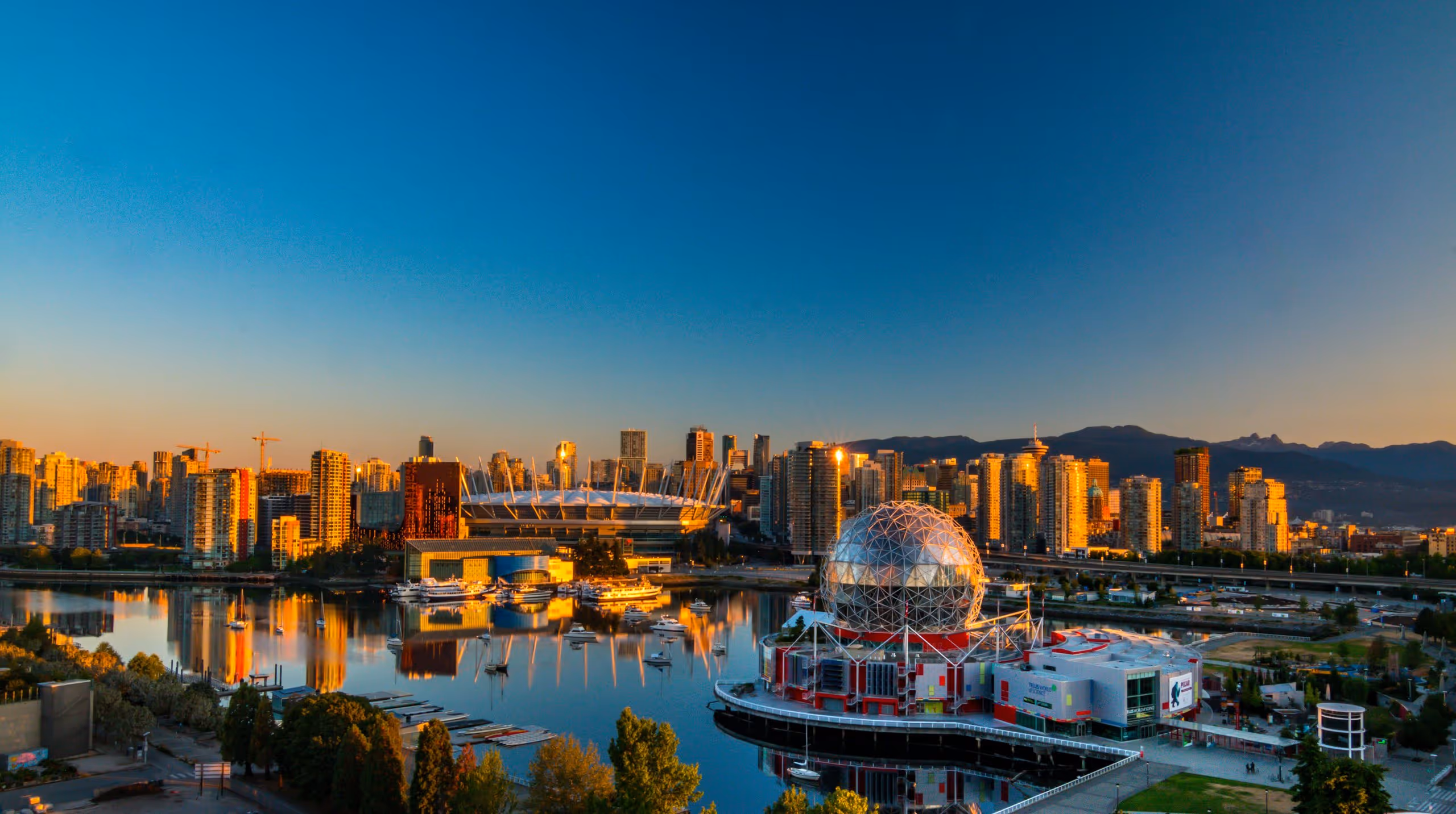 Panoramic view of a city skyline with modern buildings, a stadium, and a geodesic dome by calm water reflecting the sunset light.