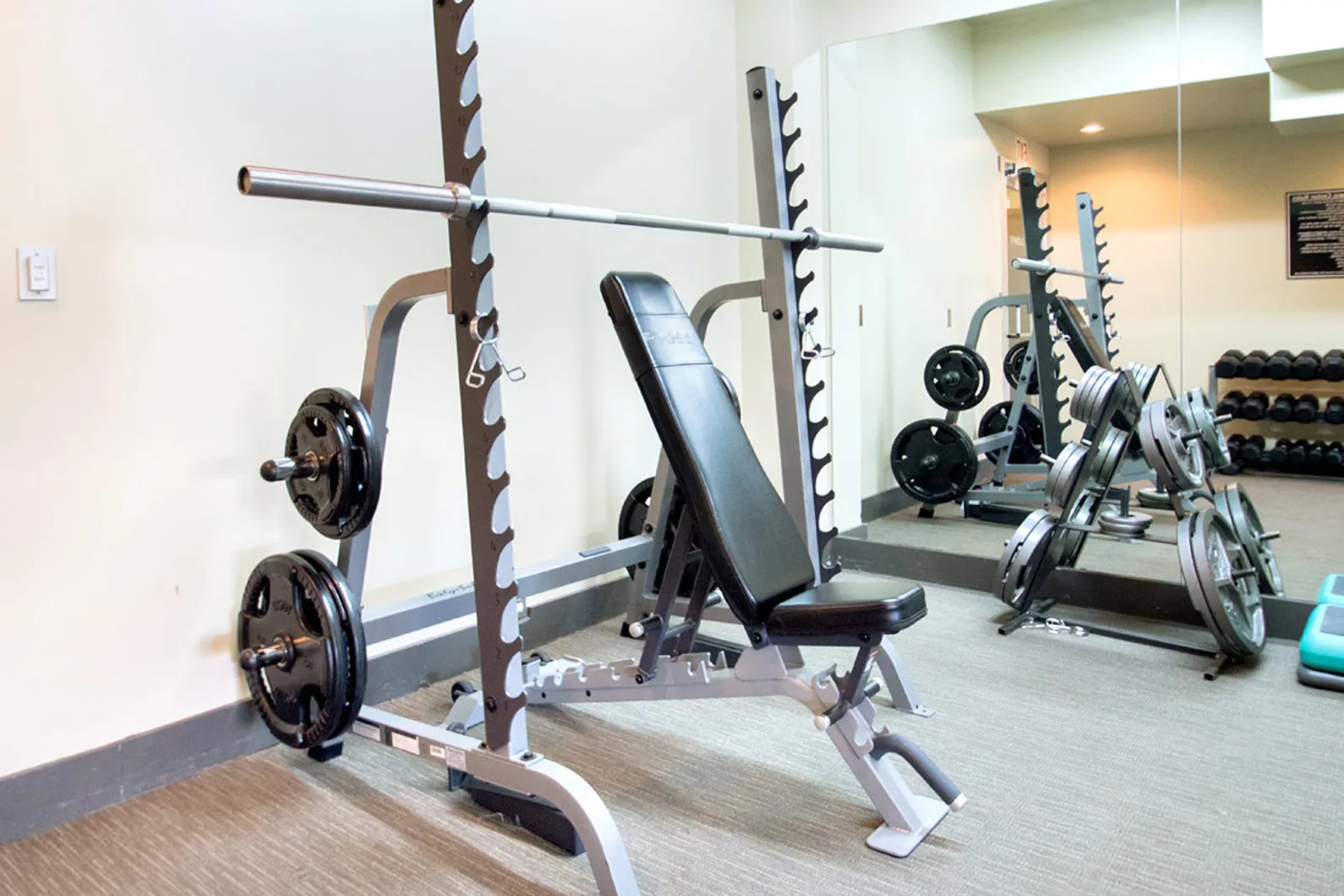 Incline bench press setup with barbell, weight plates, and a large wall mirror in a gym room.