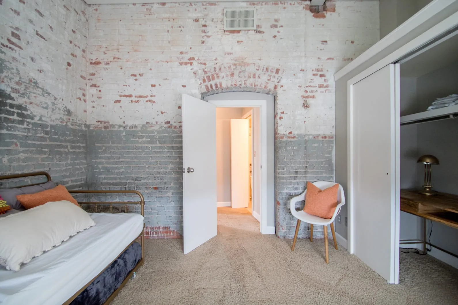 Room with exposed brick walls painted white and gray, beige carpet, a brass bed with pillows, a white chair with an orange pillow, and an open door leading to a hallway.