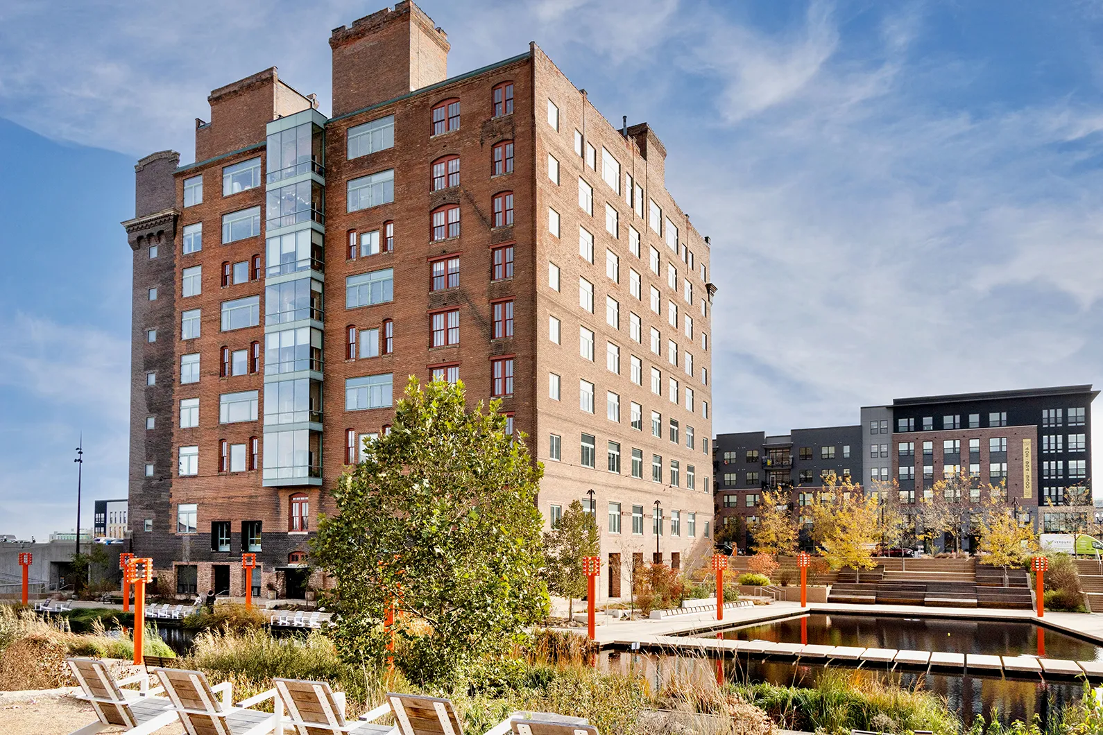 Modern brick apartment building next to a landscaped pond with wooden chairs and walking paths under a blue sky.