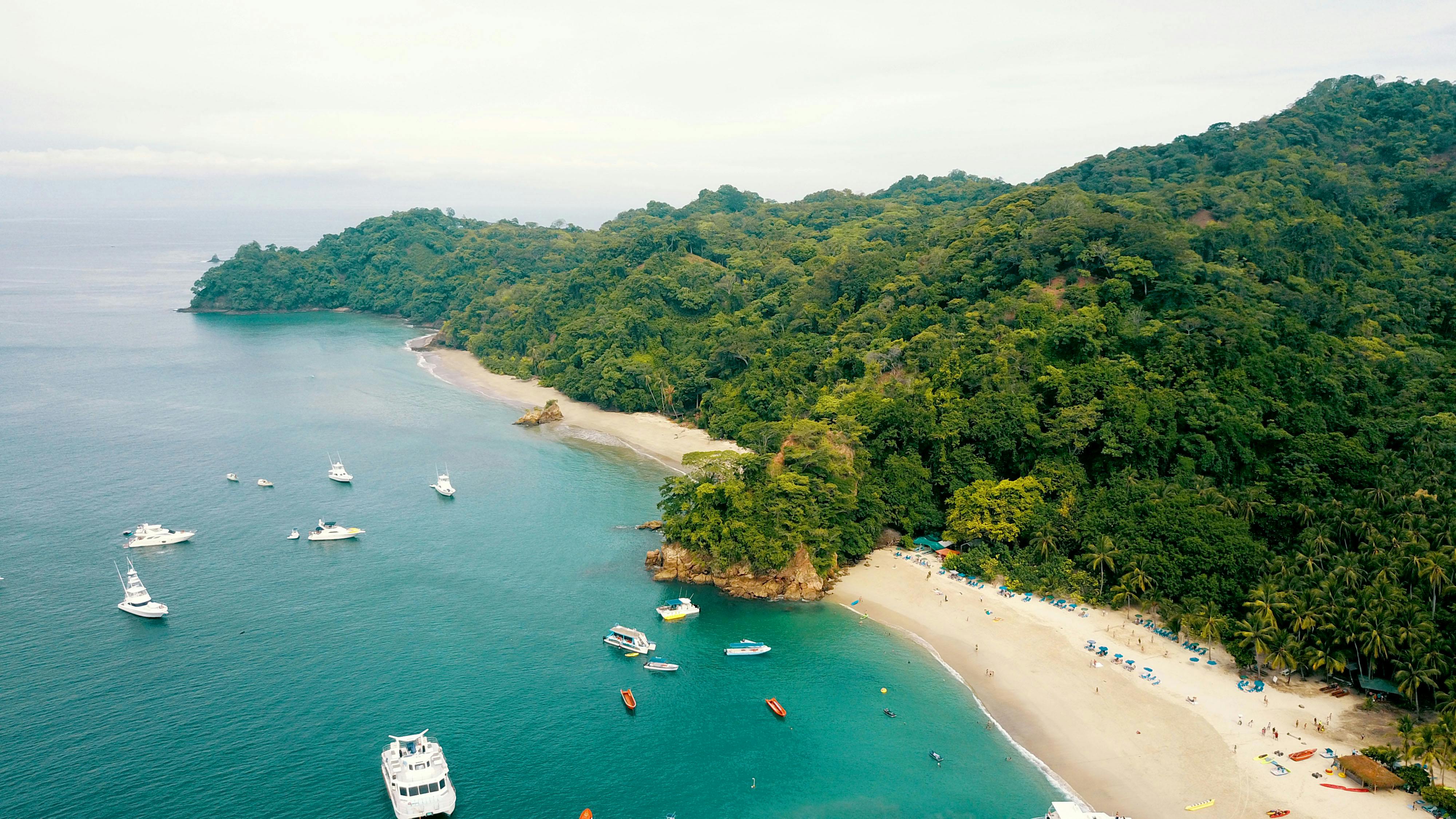 Aerial view of a turquoise bay with several boats anchored near a sandy beach bordered by lush green forest.