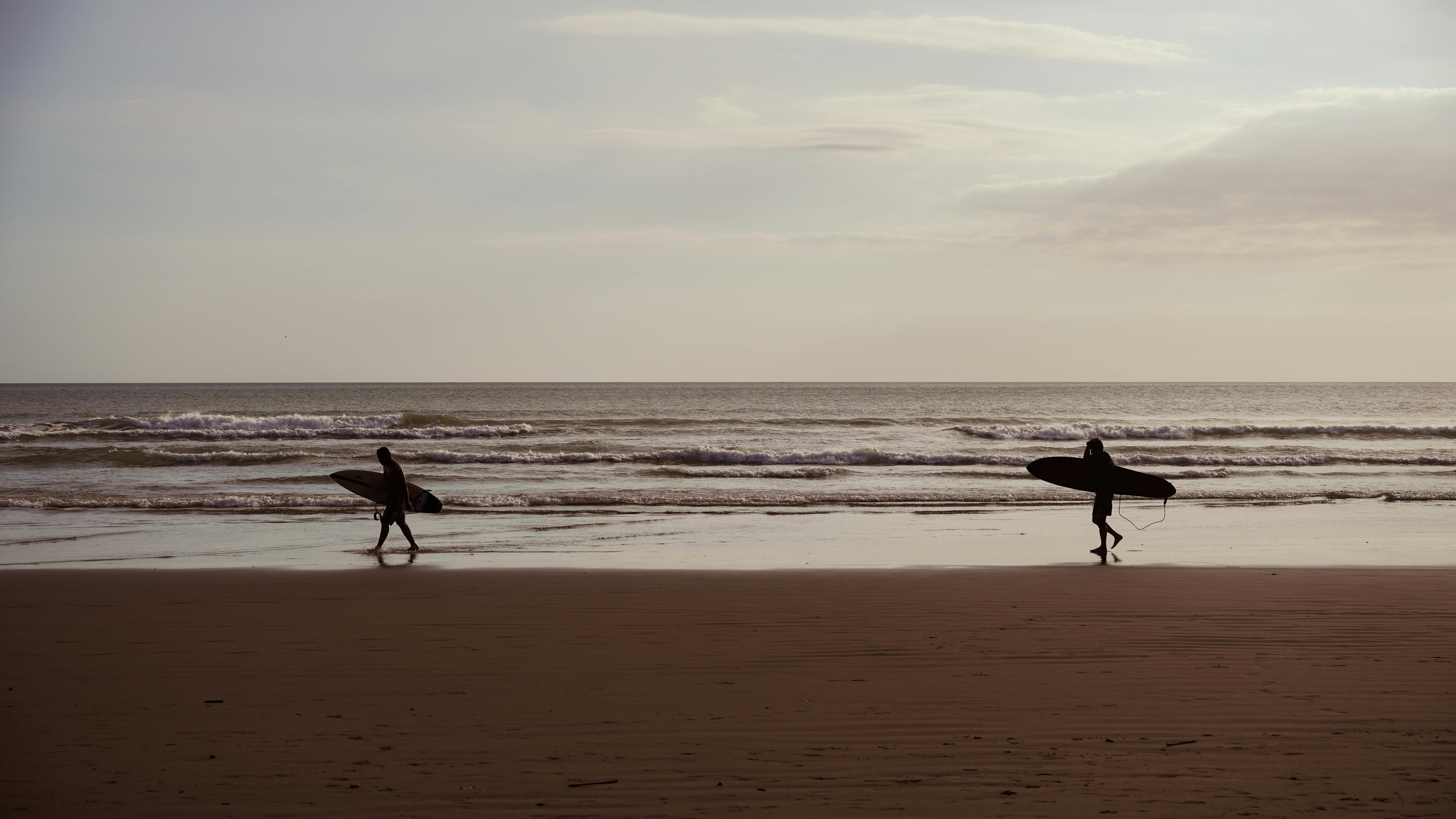 Silhouettes of two surfers walking on a beach carrying surfboards at sunset with gentle ocean waves in the background.