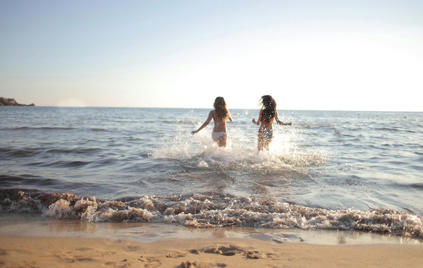 Two women in swimsuits running into the ocean water on a sandy beach at sunset.