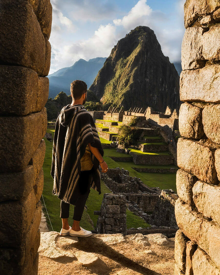 Man in traditional poncho standing between stone walls looking at Machu Picchu ruins and mountain in the background.