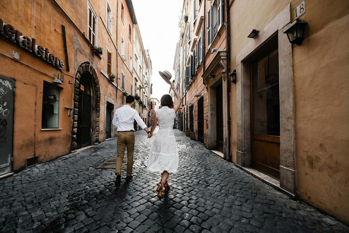 Couple holding hands walking down a cobblestone street lined with old buildings, with the woman raising a hat in the air.