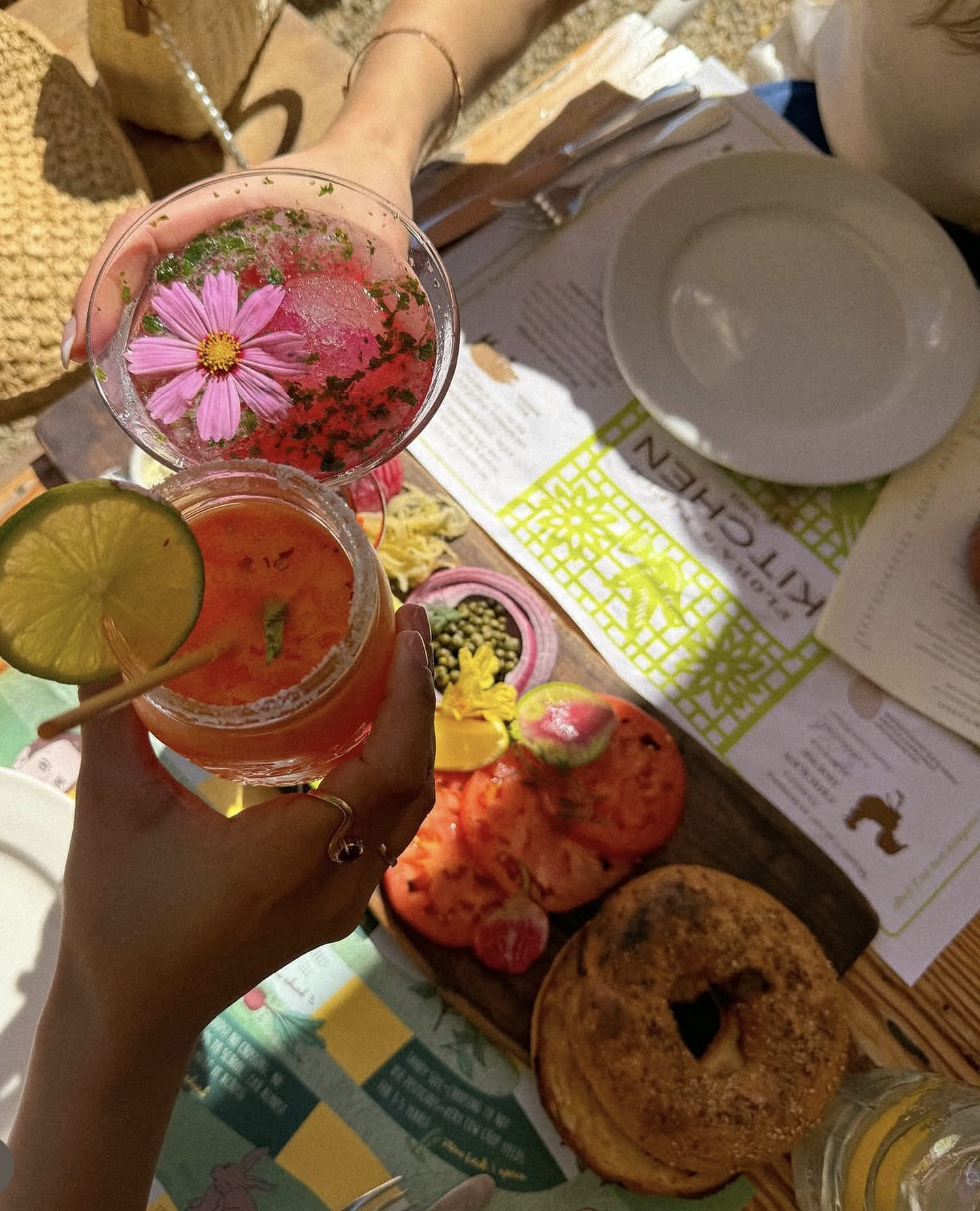 Two hands holding colorful cocktails above a table with sliced tomatoes, bagel, and menu on a wooden surface.