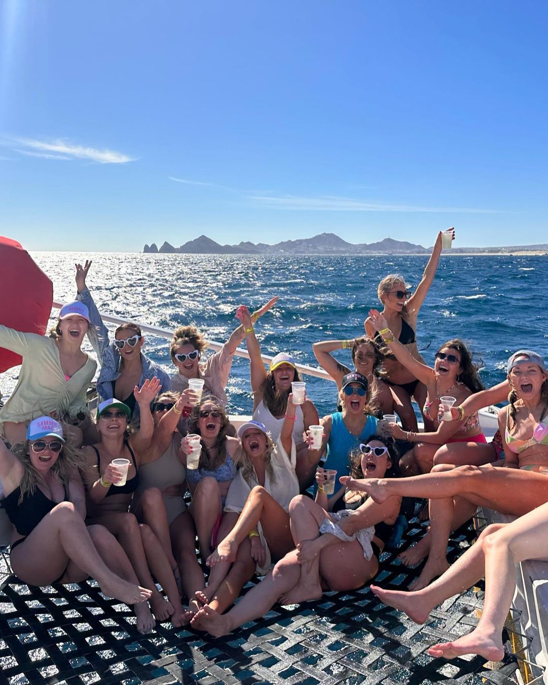 Group of women in swimsuits and sunglasses celebrating with drinks on a boat over ocean water with mountains in the background.