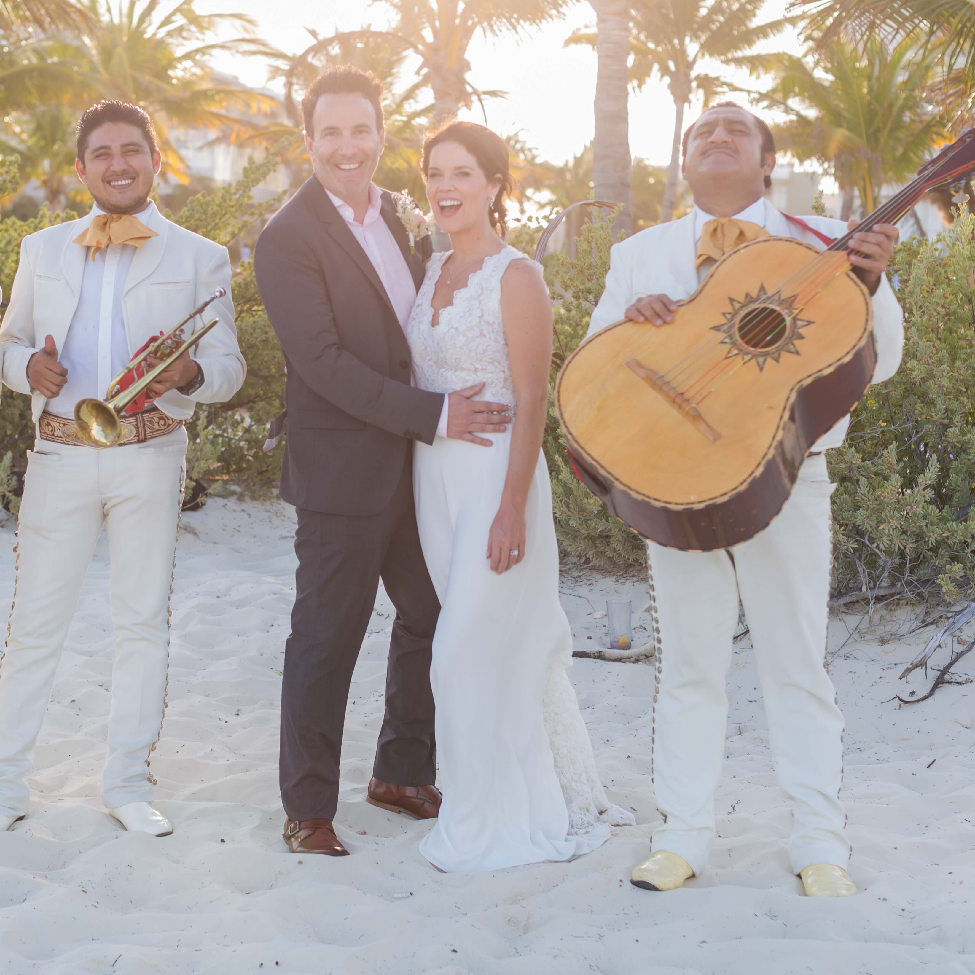 Newlywed couple standing on a sandy beach surrounded by two musicians in white suits, one holding a trumpet and the other a guitar.