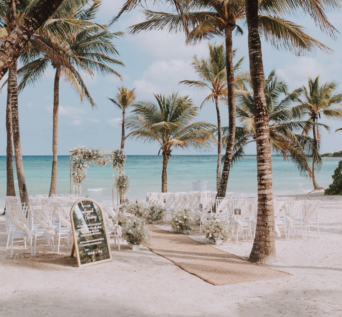 Outdoor beach wedding setup with white chairs, floral arrangements, and a wooden arch against a backdrop of palm trees and turquoise ocean.