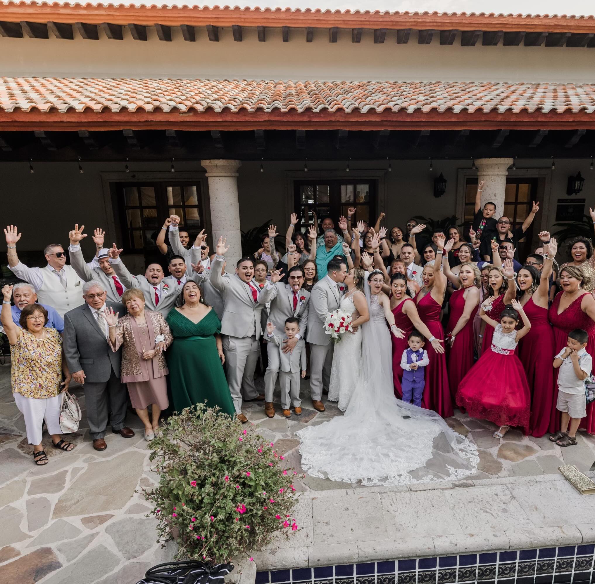 Large wedding group photo with bride and groom kissing at the center, surrounded by bridesmaids in red dresses, groomsmen in gray suits, children, and guests all raising their hands in celebration.