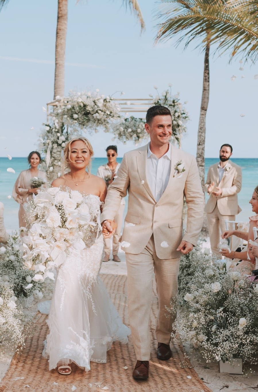 Bride and groom holding hands and walking down the aisle outdoors by the beach, surrounded by wedding guests and floral decorations.