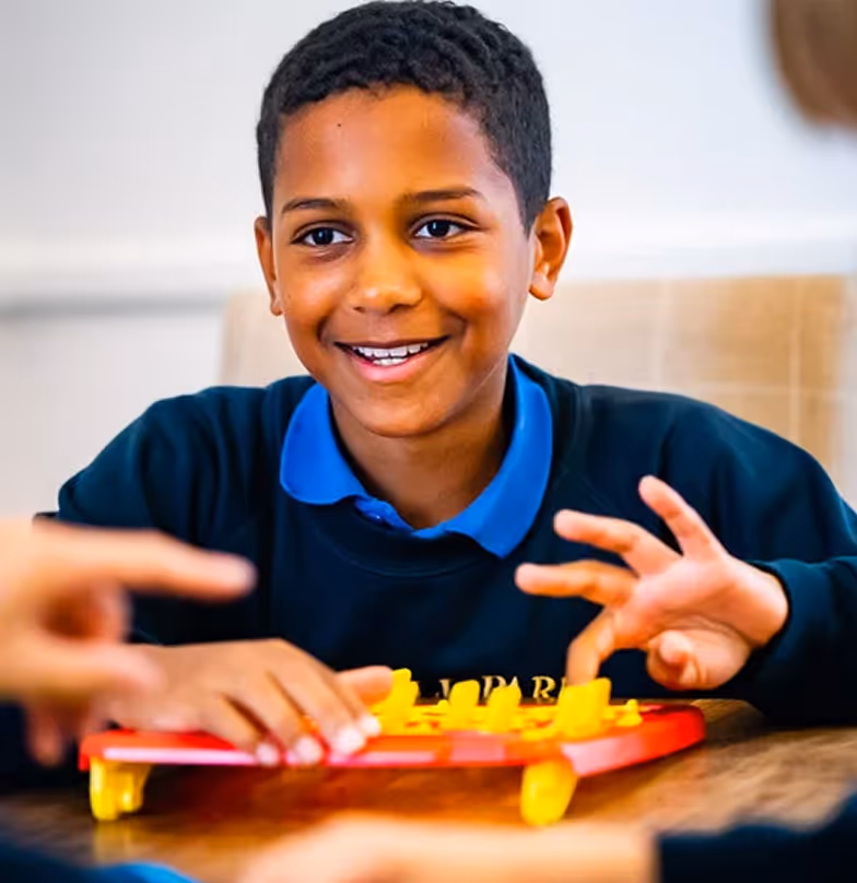 Smiling boy in blue school uniform playing a yellow and red board game at a table.
