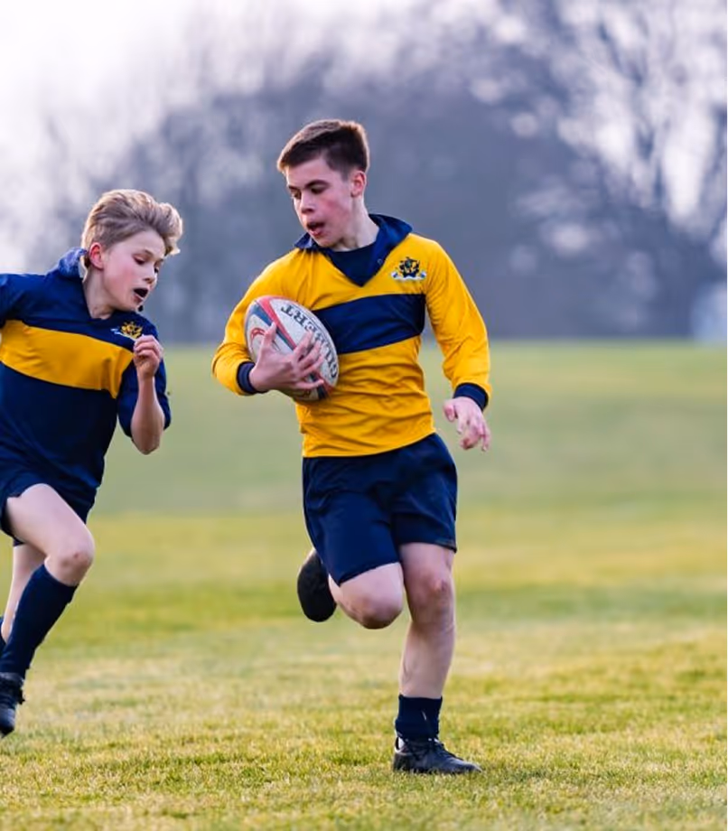 Two boys playing rugby on grass, one running with the ball while the other chases.