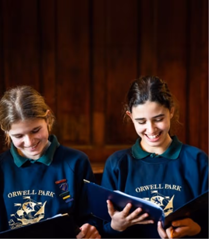 Two smiling students wearing Orwell Park sweatshirts reading from folders indoors.