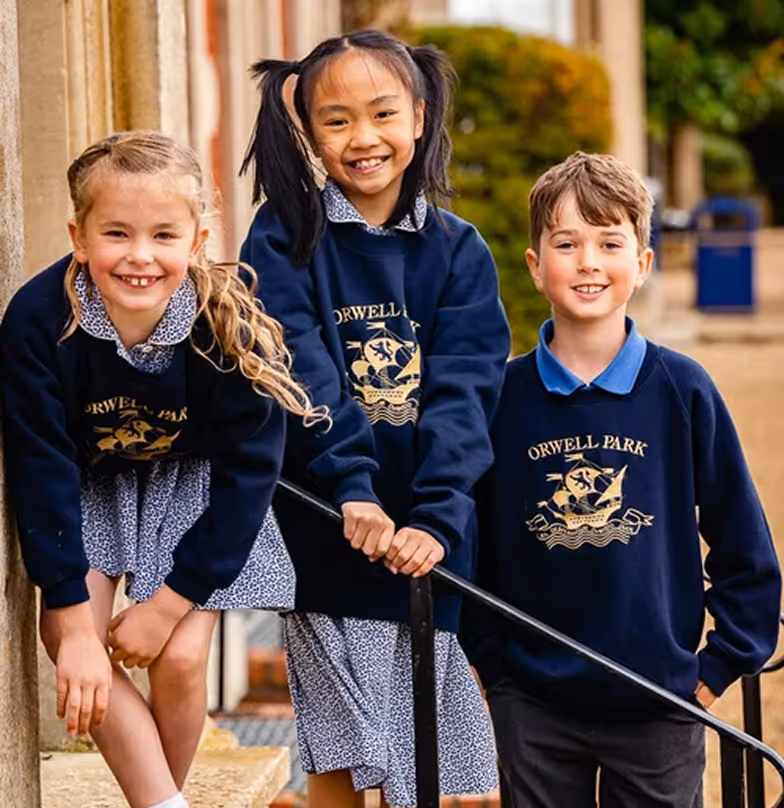 Three smiling children wearing Orwell Park school uniforms standing outside near a handrail.