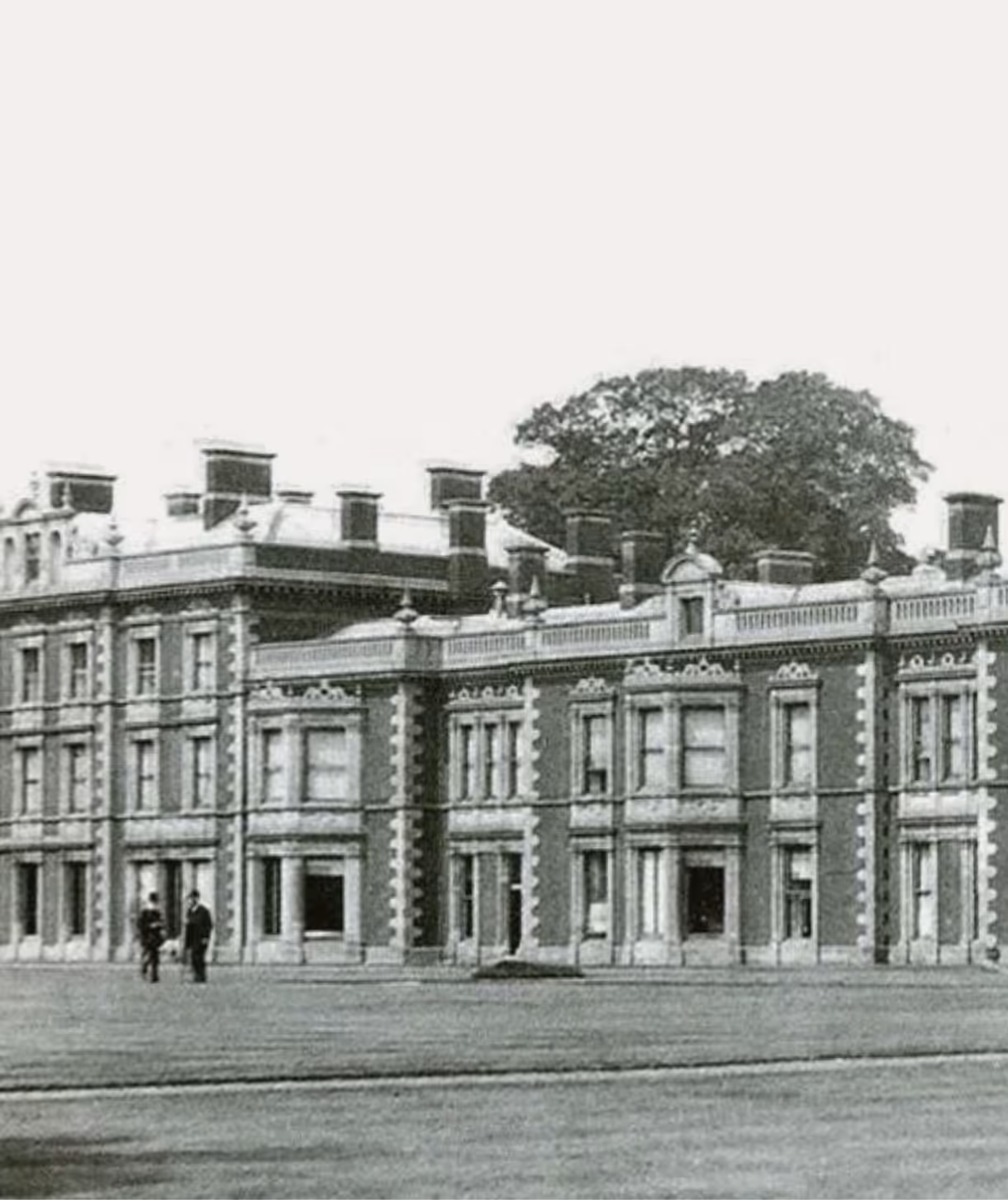 Black and white photo of a large historical brick building with multiple chimneys and two people standing on the lawn in front.