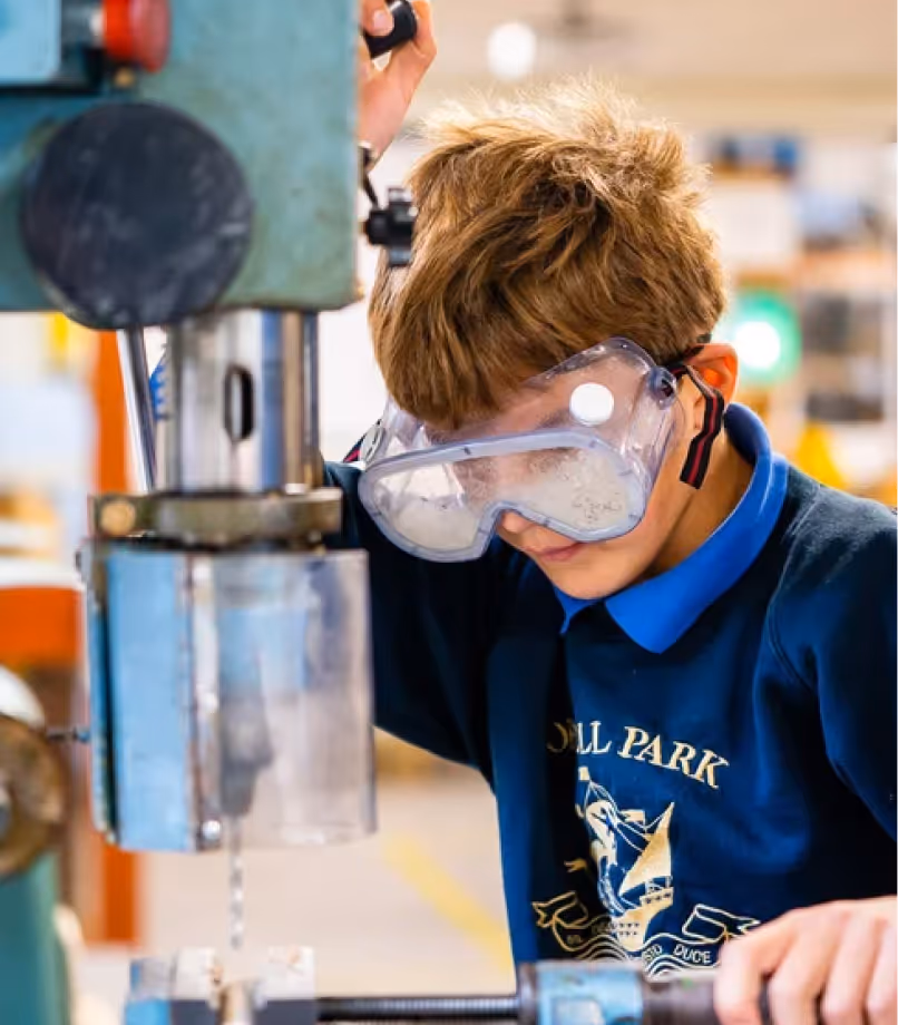 Young boy wearing protective goggles operating a drill press machine in a workshop.