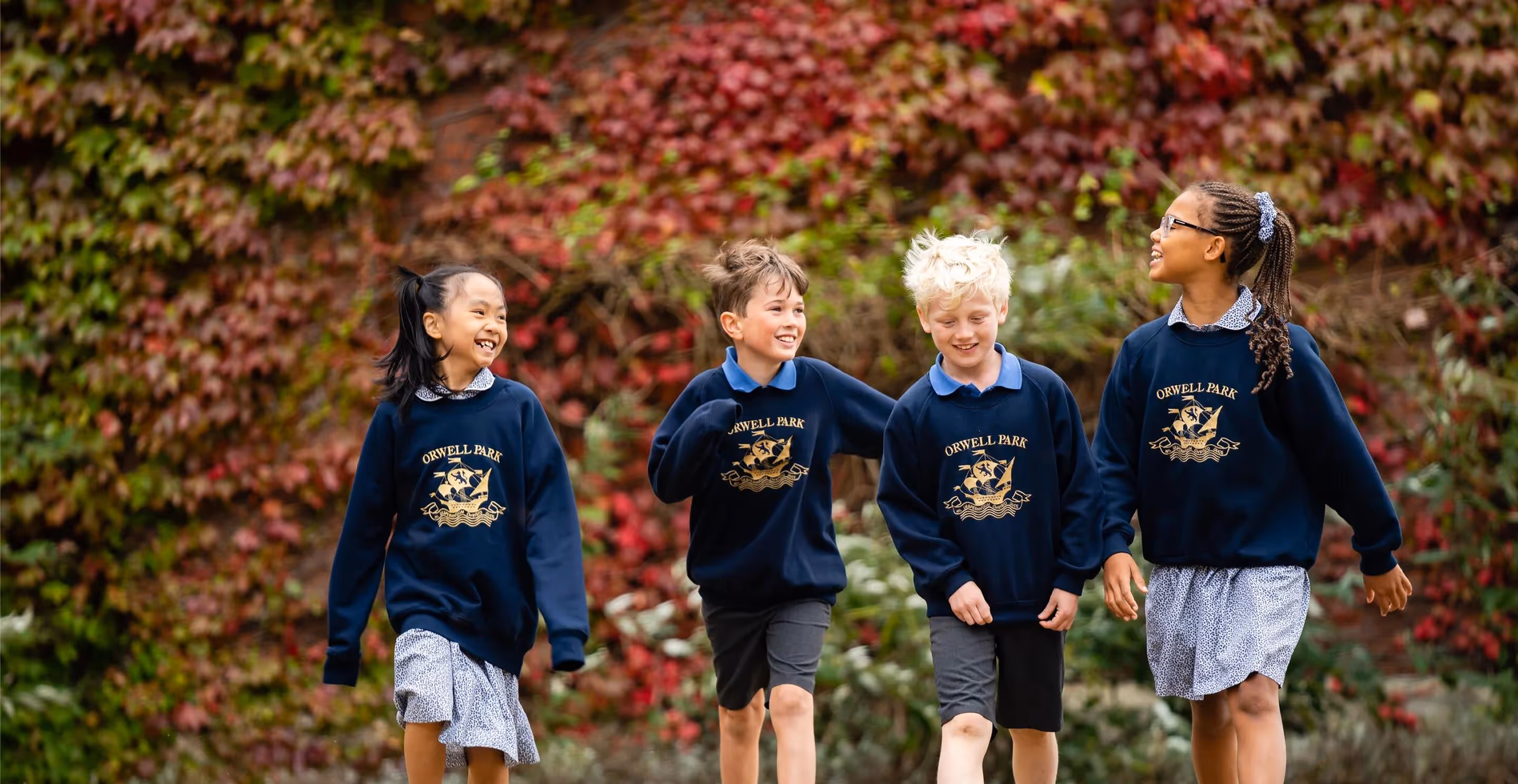 Four children walking and smiling outdoors wearing Orwell Park School navy sweatshirts in front of a leafy wall.