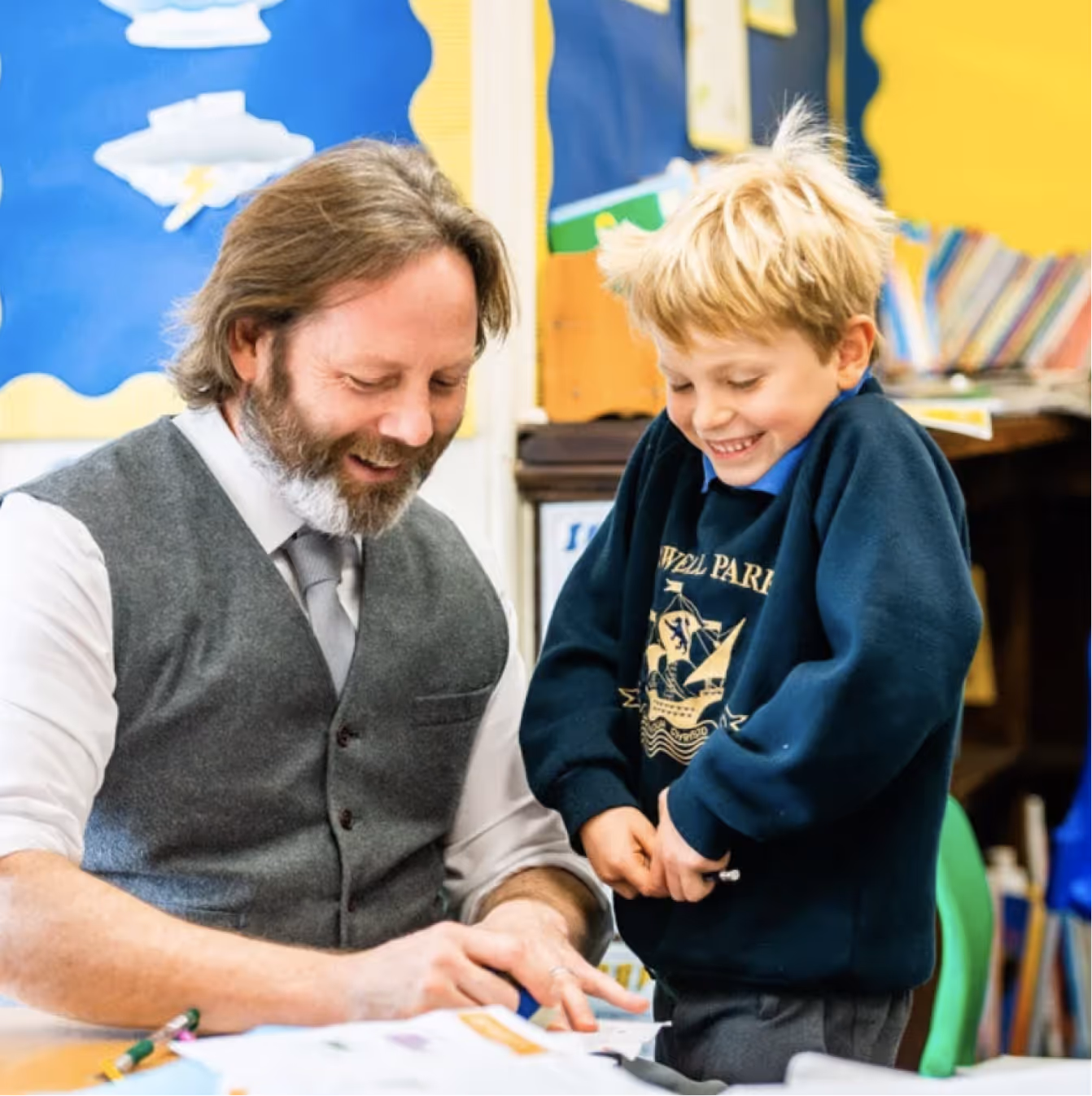 Smiling teacher assisting a young boy in a classroom with colorful bulletin boards and books in the background.
