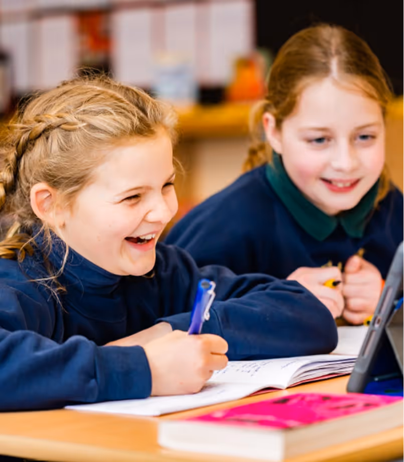 Two young girls in school uniforms smiling and working together on a tablet at a desk with notebooks.