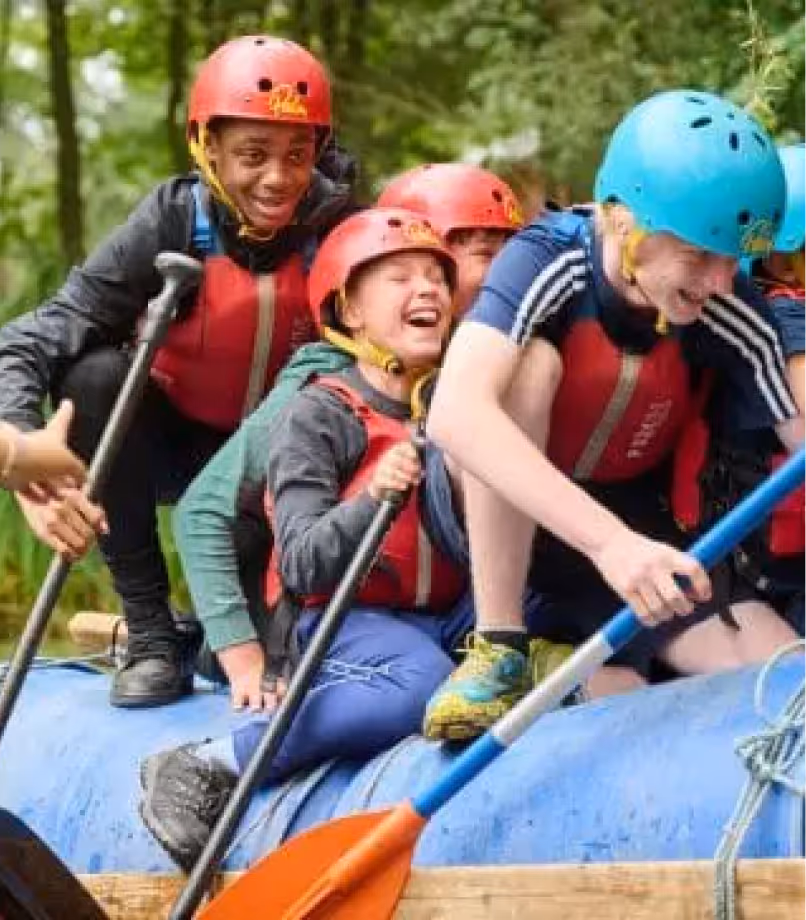Group of young people wearing helmets and life jackets paddling together on a blue raft in a wooded area.