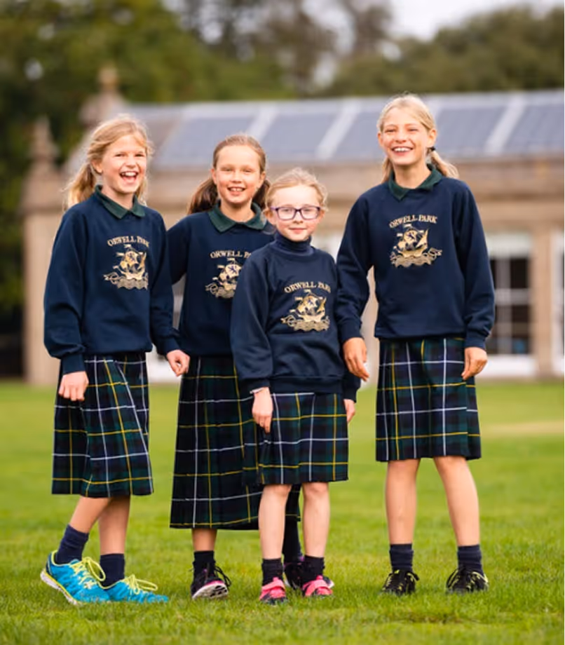 Four schoolgirls in navy sweaters and plaid skirts smiling while standing on grass outdoors.